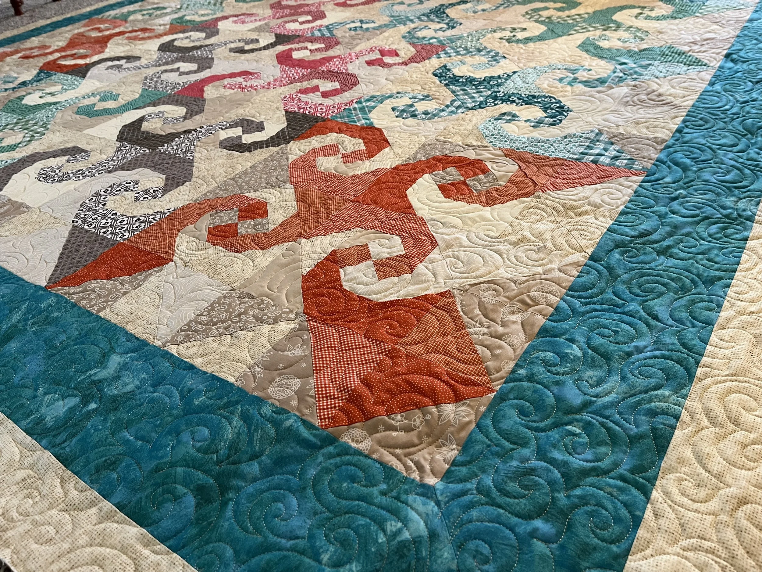 Close-up of a colorful quilt with intricate quilting patterns. The quilt features various fabric patterns in red, teal, gray, cream, and black, arranged in geometric blocks with a teal border.