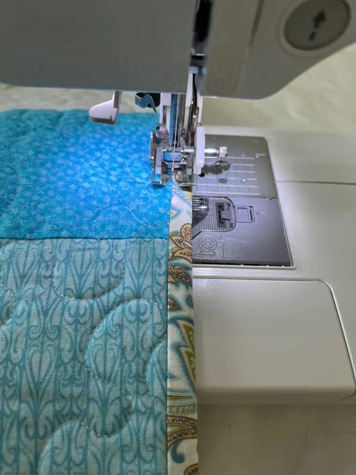 Close-up view of a sewing machine stitching a quilt with a blue patterned fabric on the left and a multicolored patterned fabric on the right.