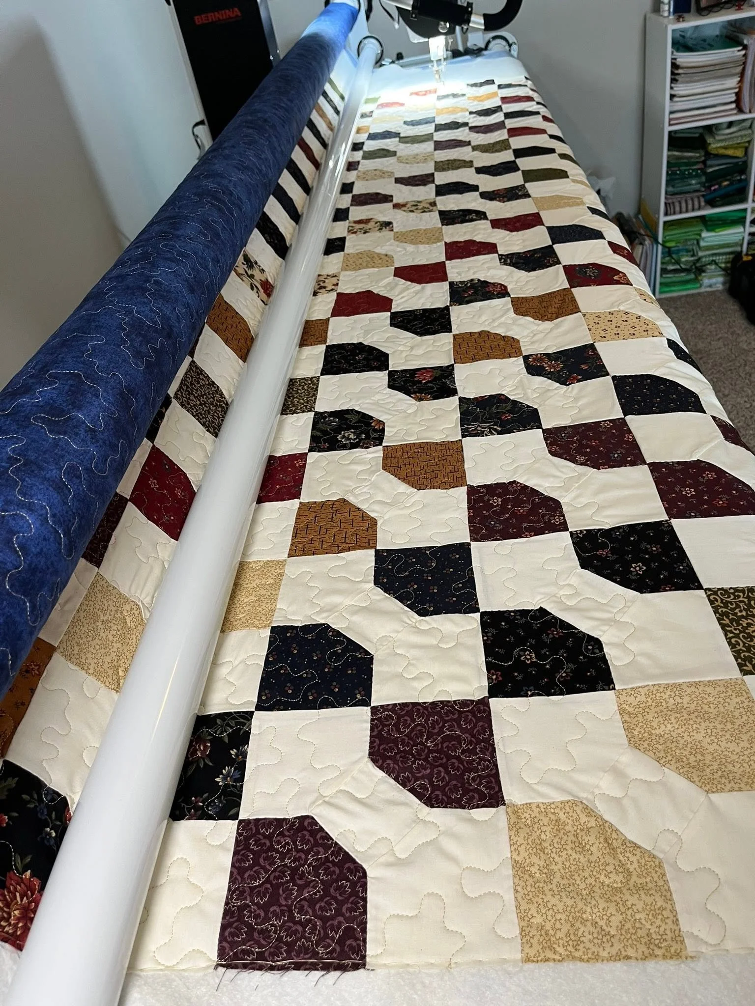 A long sewing table with a colorful patchwork quilt in progress, a large blue fabric rolled on a machine, white piping, and a shelf filled with fabric and other supplies in the background.