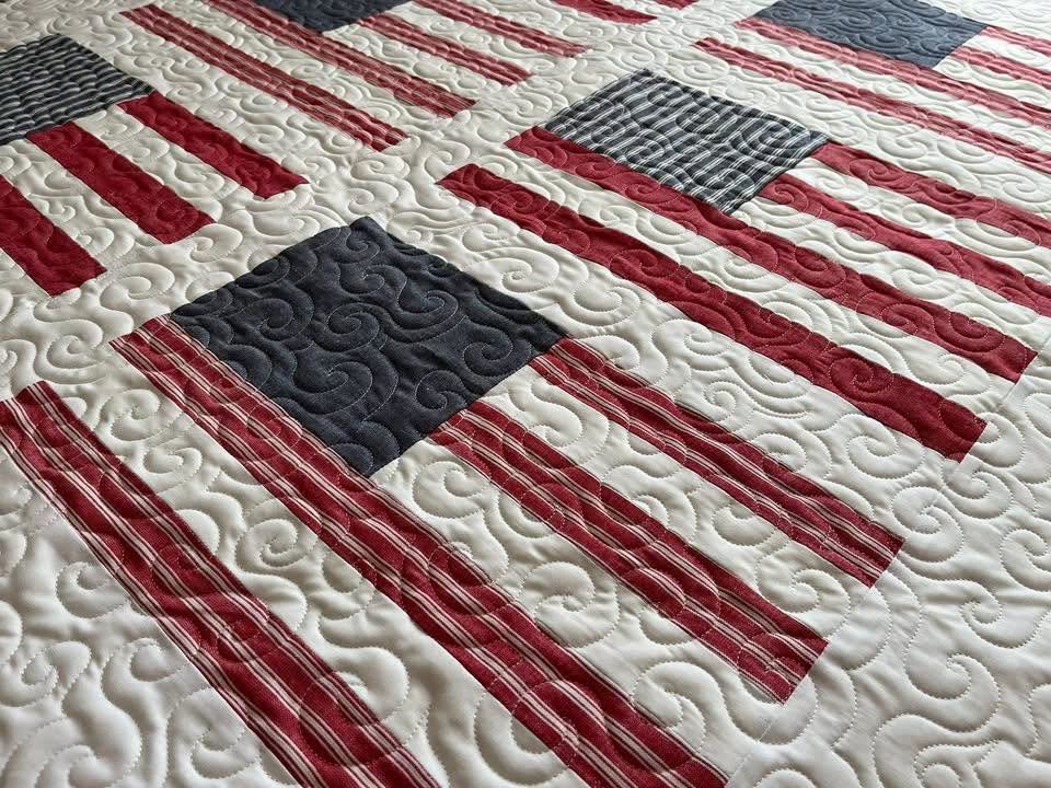 A quilt featuring a pattern of red, white, and blue stripes with some solid and plaid squares, reminiscent of the American flag.