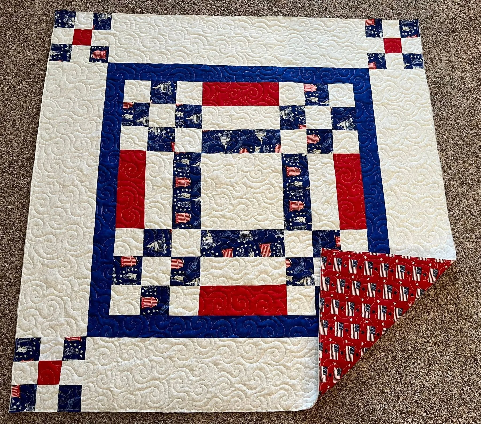 A quilt with a patriotic theme featuring white, red, and blue fabric with stars, stripes, and American flag patterns, laid out on a beige carpet.