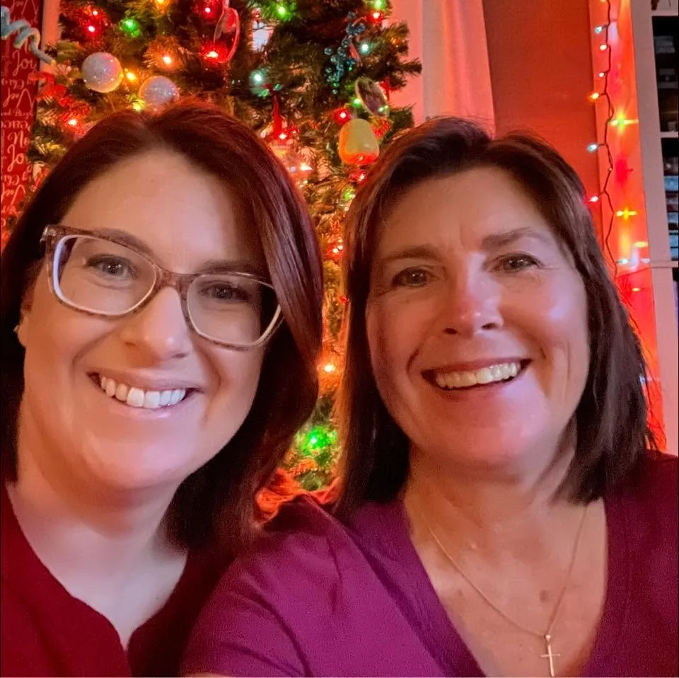 Two women smiling in front of a decorated Christmas tree with colorful lights.
