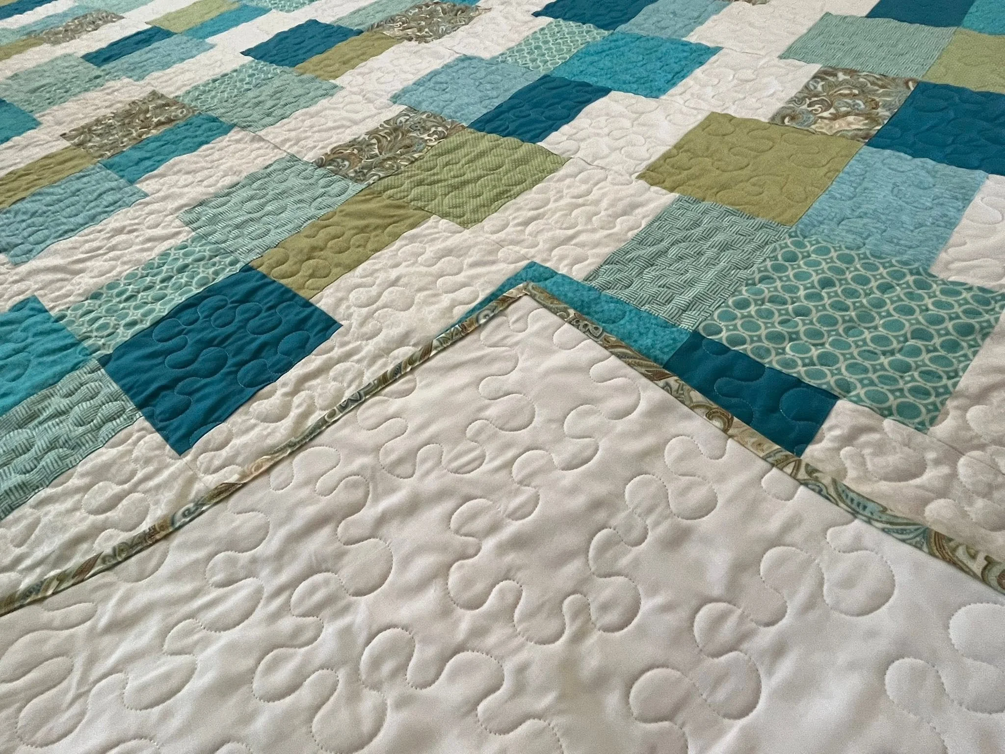 Close-up of a quilt with various blue, green, and white fabric patches arranged in a pattern, with some patches featuring swirling and geometric designs.