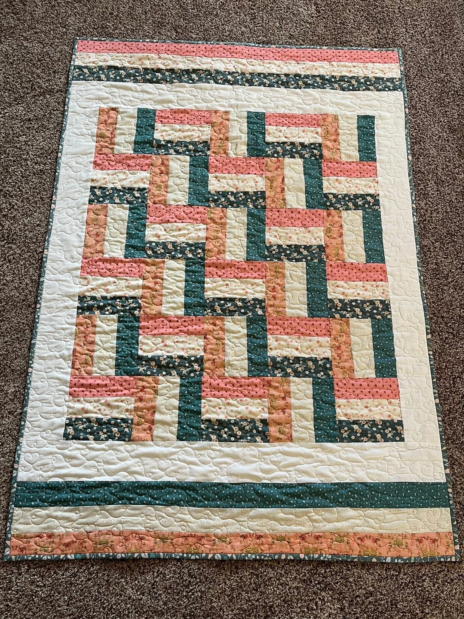 A rectangular patchwork quilt with a floral and dotted fabric pattern, in pink, white, and dark blue colors, placed on a brown carpeted surface.