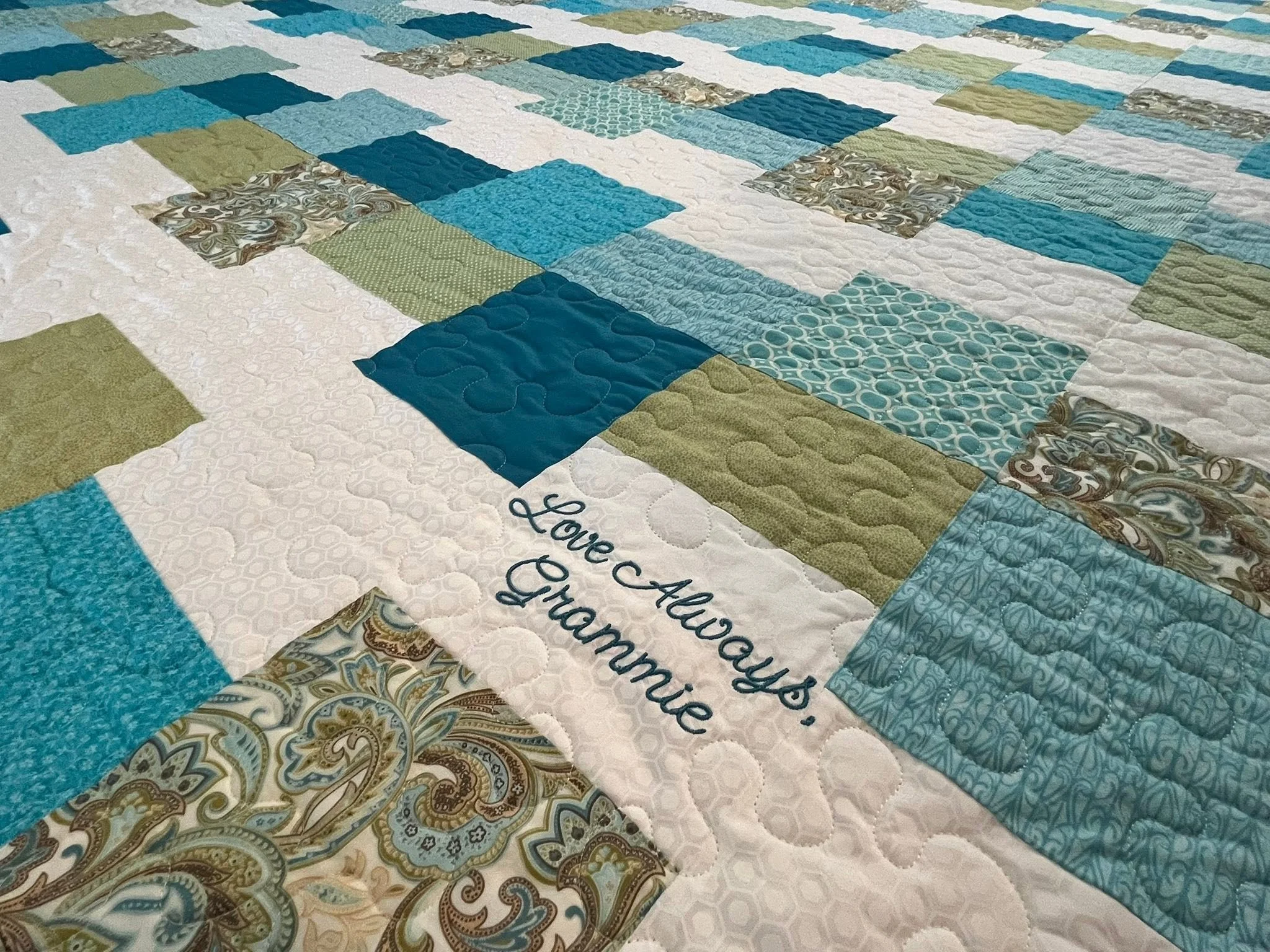 A close-up of a handmade quilt with various patterned fabric squares in shades of blue, green, brown, and white, arranged in a grid pattern.
