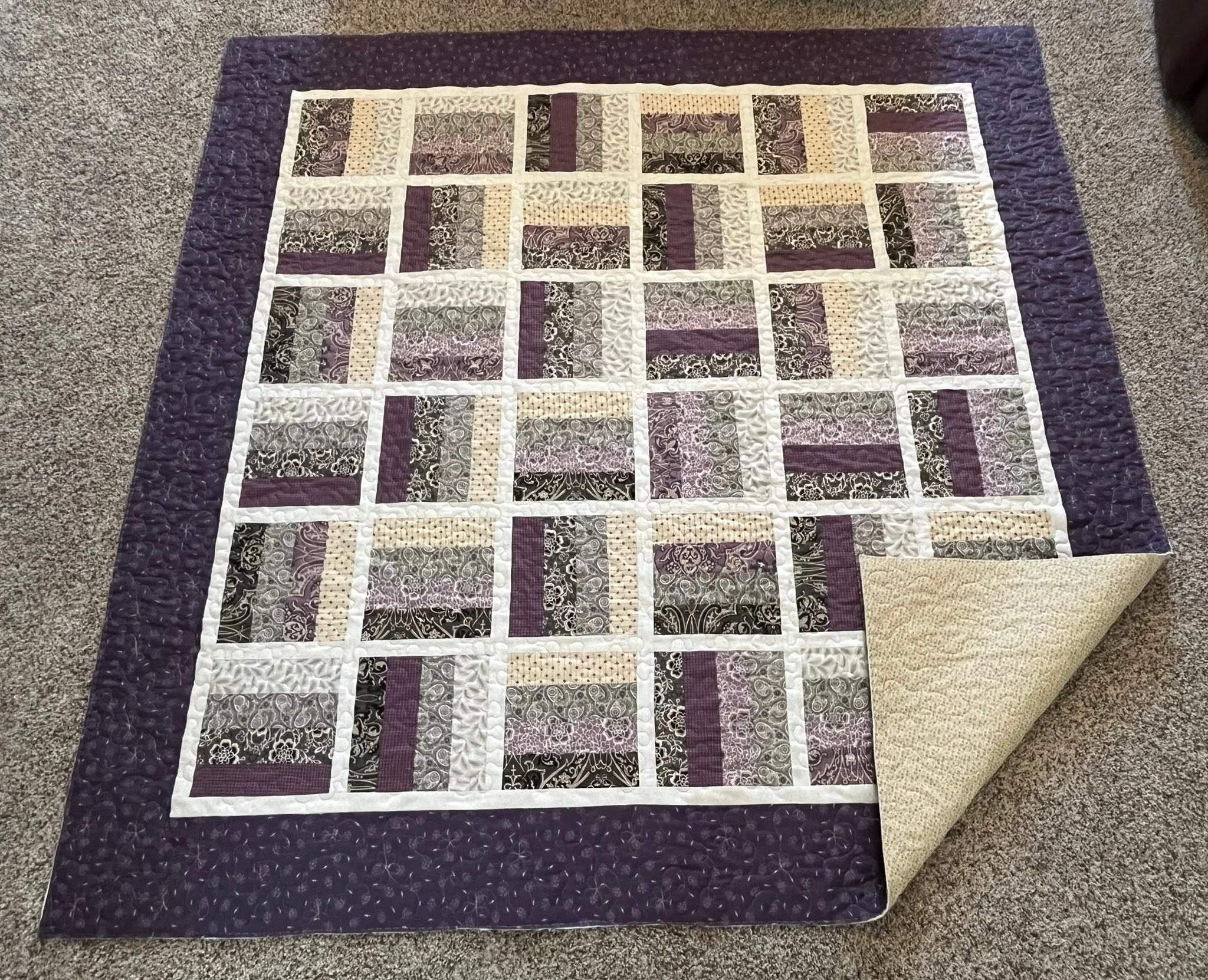 A quilt with a patchwork design, primarily in shades of purple, cream, and black, laid out flat on a carpeted floor.