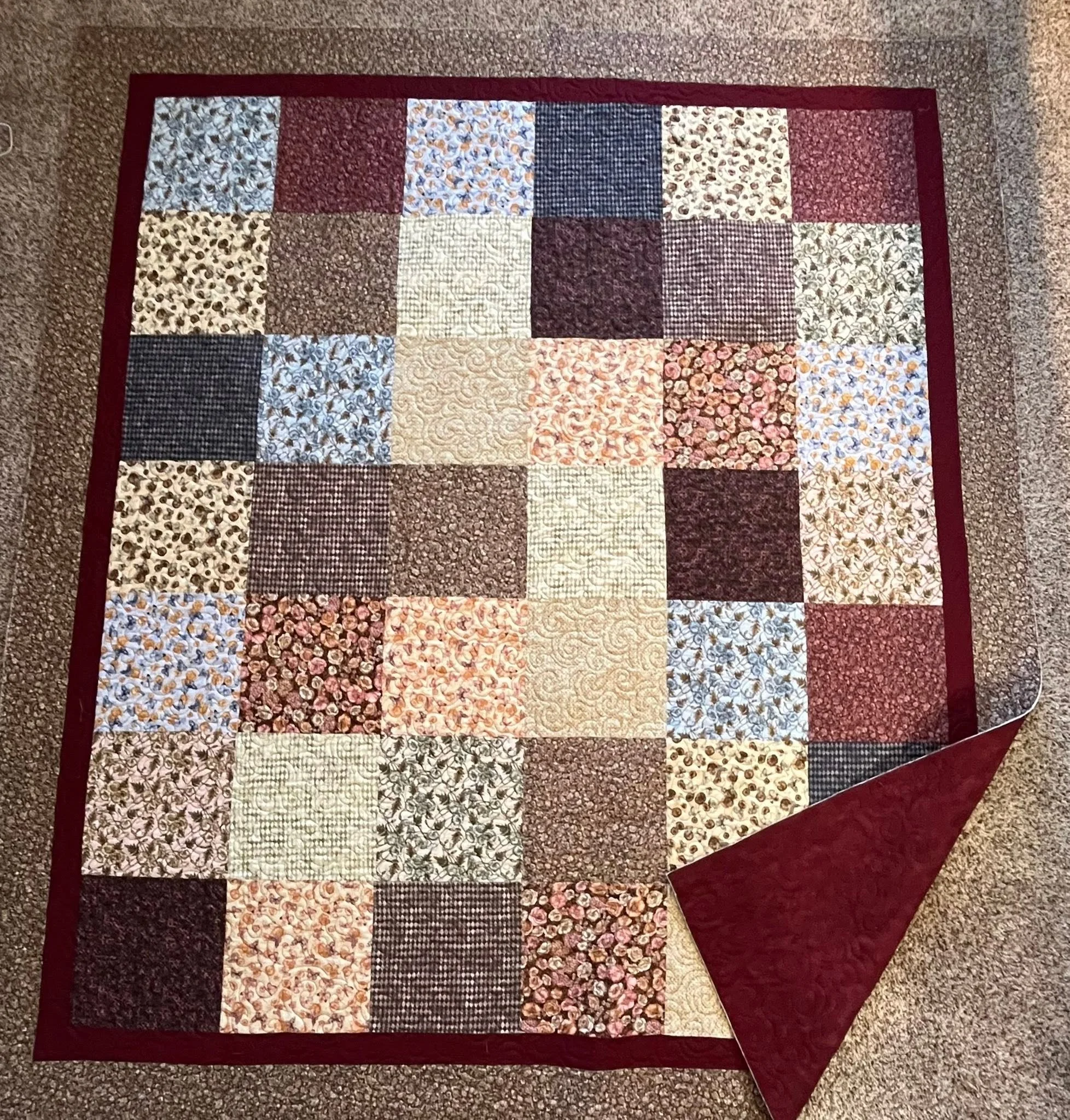 A quilt on a floor featuring a patchwork of various floral, checkered, and patterned fabric squares, bordered with a red border and a red backing.