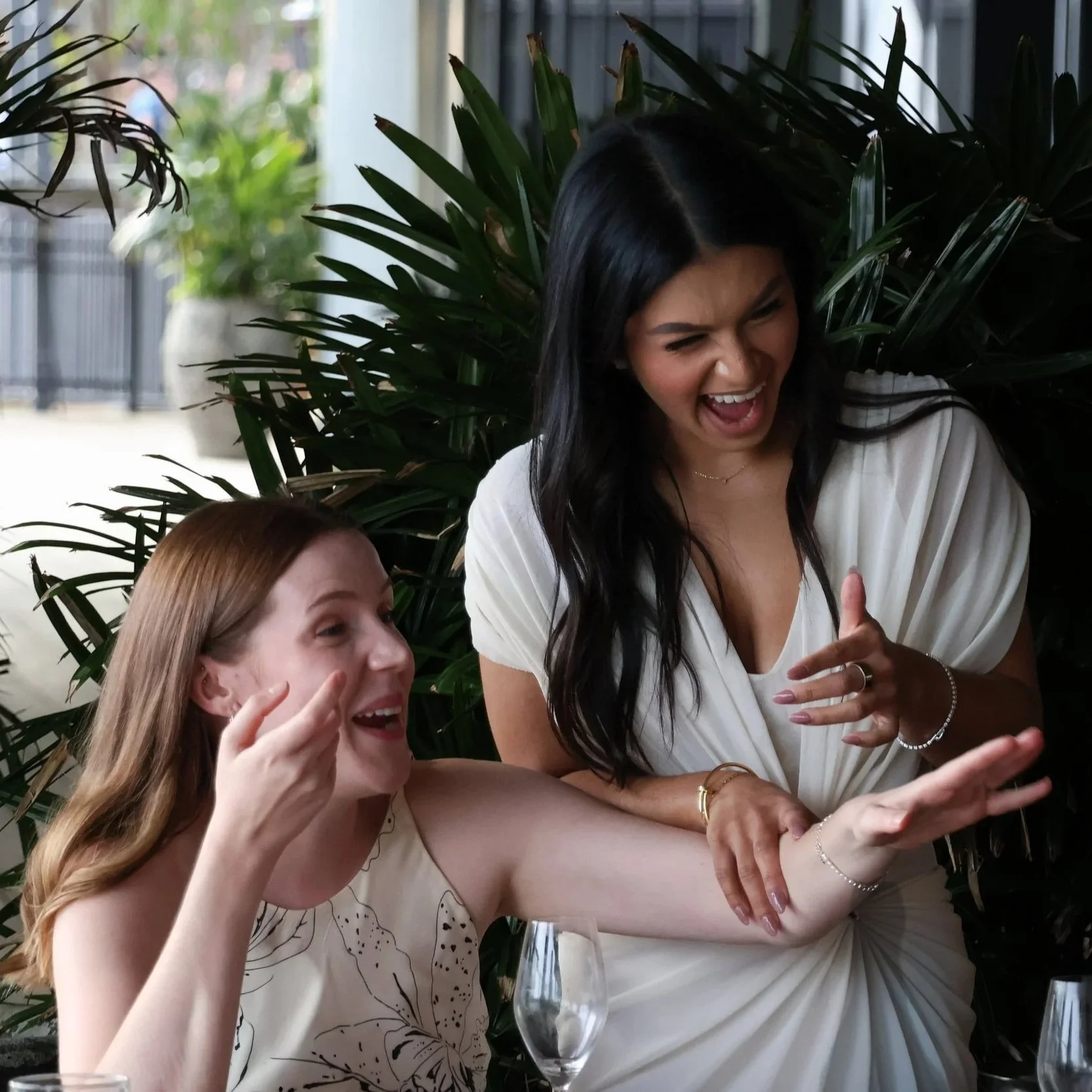 Two women laughing and smiling, one with long blonde hair and the other with long black hair, sitting in front of green plants indoors.