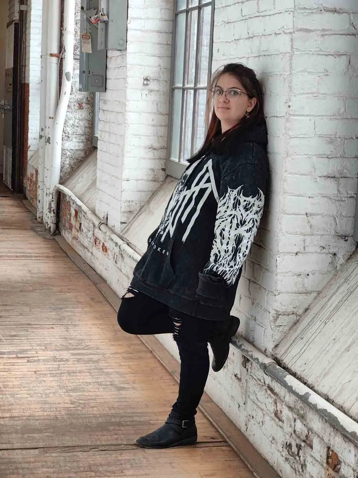 A young woman with glasses and long hair leaning against a white brick wall, wearing a black hoodie with white graphics and ripped black jeans in an industrial-style space.