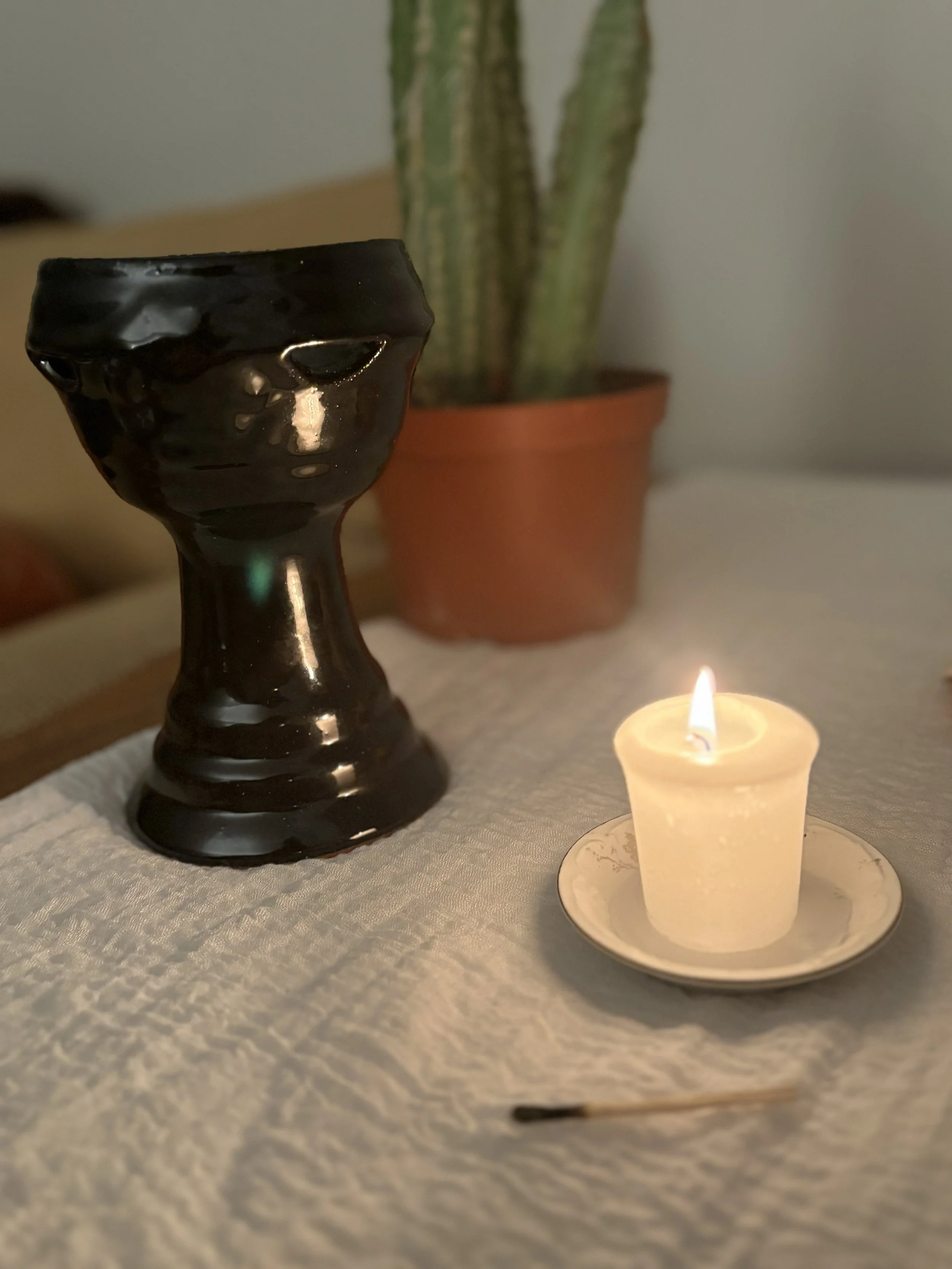 A lit candle in a small container, a black ceramic object resembling a face or mask, and a potted cactus plant in the background on a table.