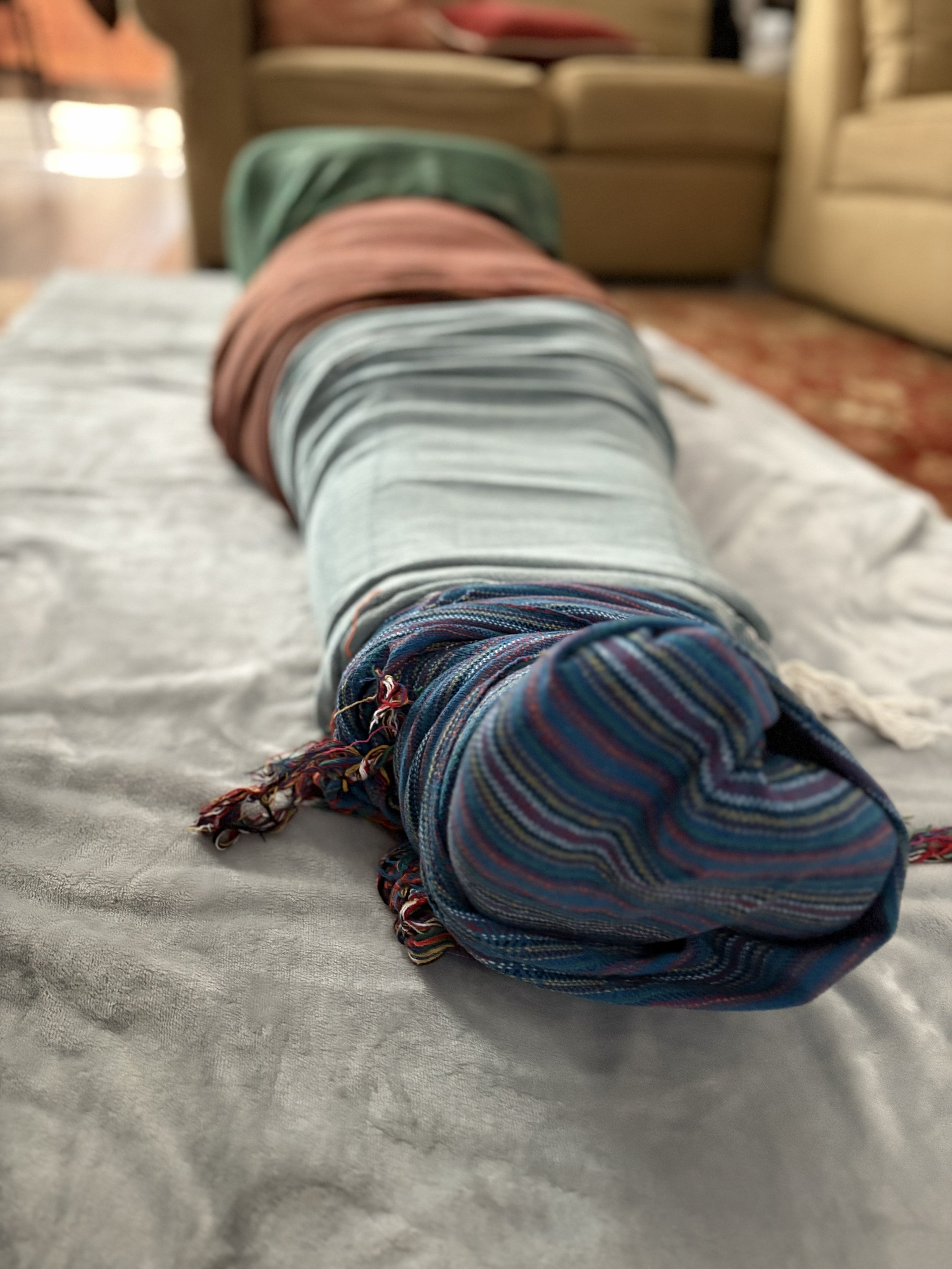 A person's legs extended straight in front, wearing colorful striped socks, resting on a soft, light-colored surface. In the background, a blurred living room with a couch and wood-paneled walls.
