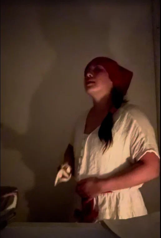 A woman wearing a red headscarf and a light-colored dress standing in a dimly lit room.