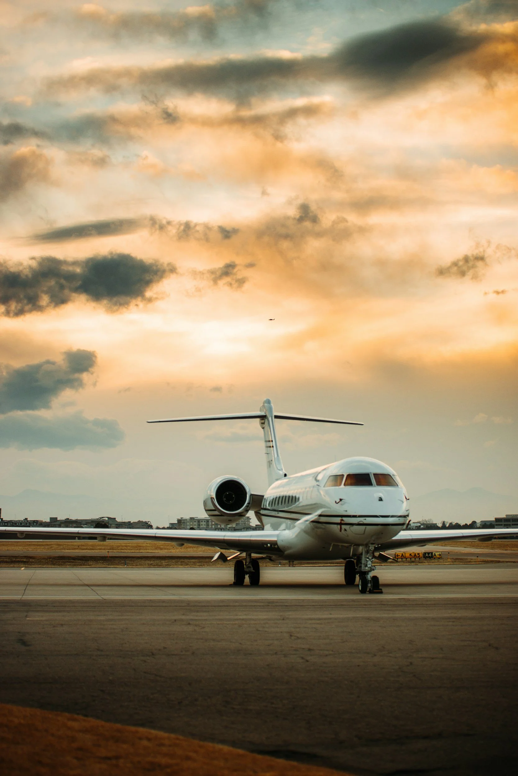A private jet parked on an airport runway during sunset, with a colorful sky filled with clouds and a small airplane in the distance.