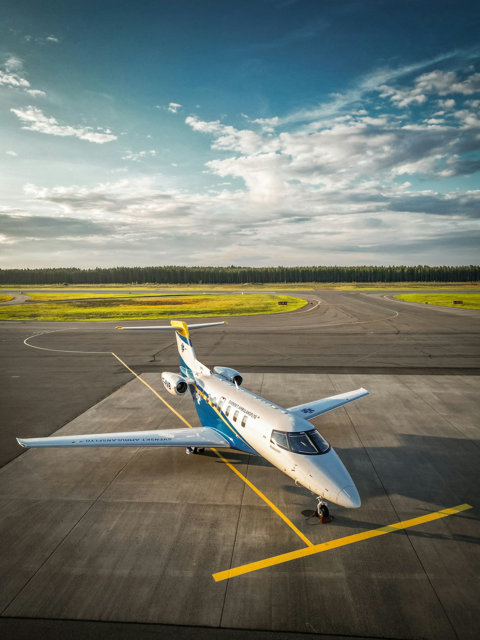 Small aircraft parked on tarmac with a yellow line, background of grassy fields and forest, cloudy sky.