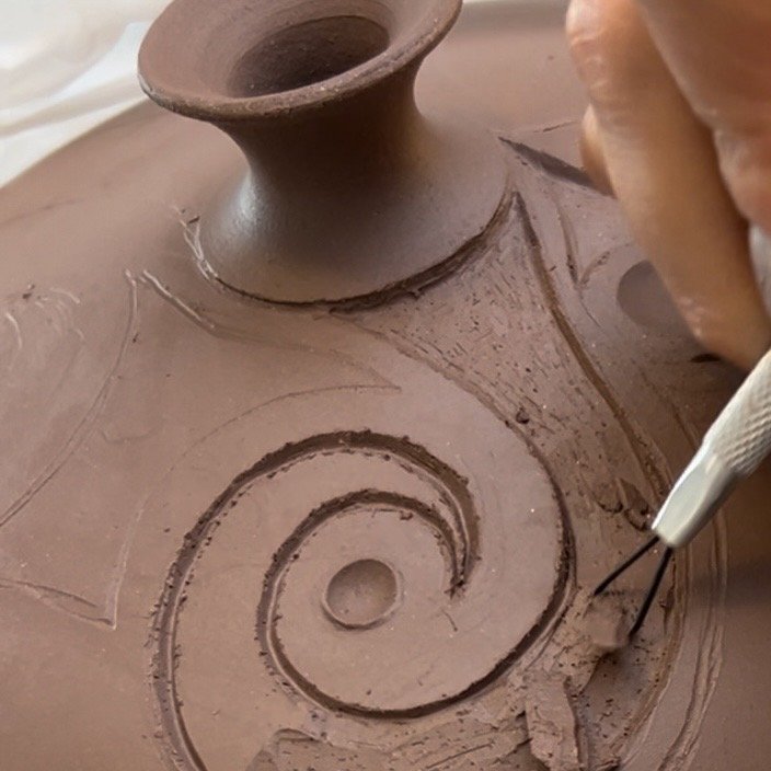 Close-up of a person carving a decorative spiral pattern into a clay pot using a small metal tool.