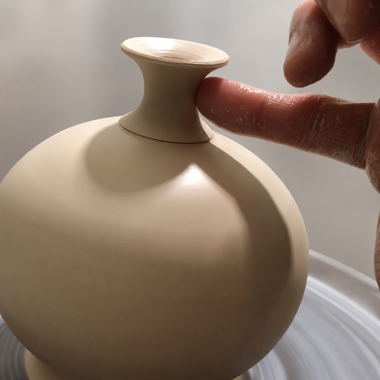 Close-up of a potter's hand with clay fingerprints touching the top of a beige, smooth, rounded ceramic vase on a pottery wheel.