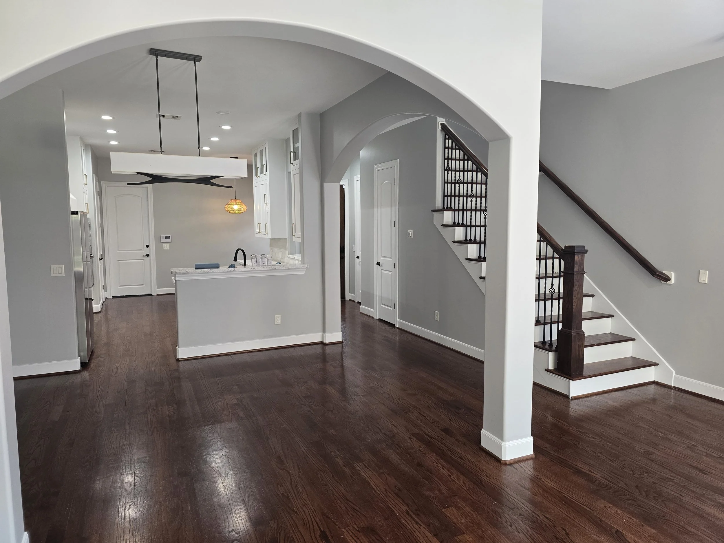 Interior view of a modern home with dark wood flooring, white walls, a staircase with dark wooden handrail and white spindles, and a kitchen area in the background with white cabinetry, a white island, and contemporary lighting fixtures.