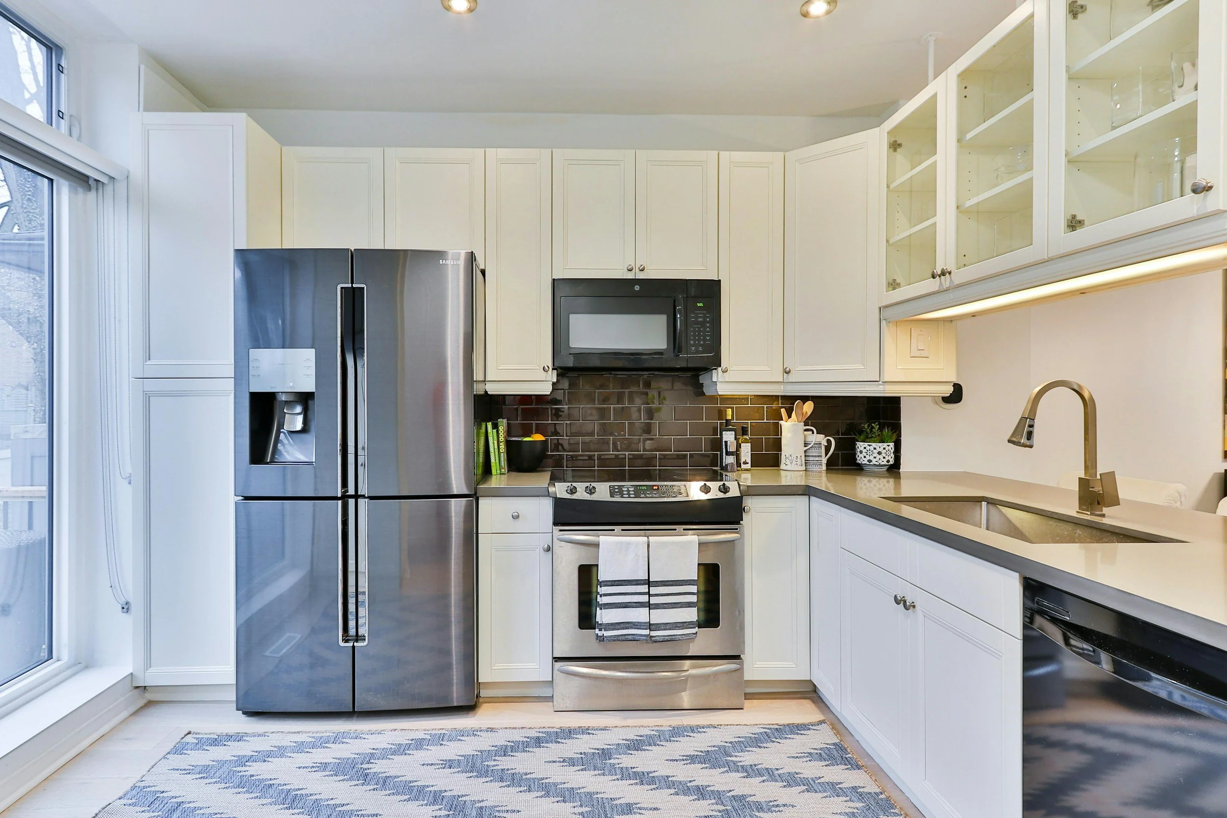 Modern kitchen with white cabinets, stainless steel refrigerator, black microwave above stove, gray countertops, and a window with a view outside. Kitchen appliance installation.