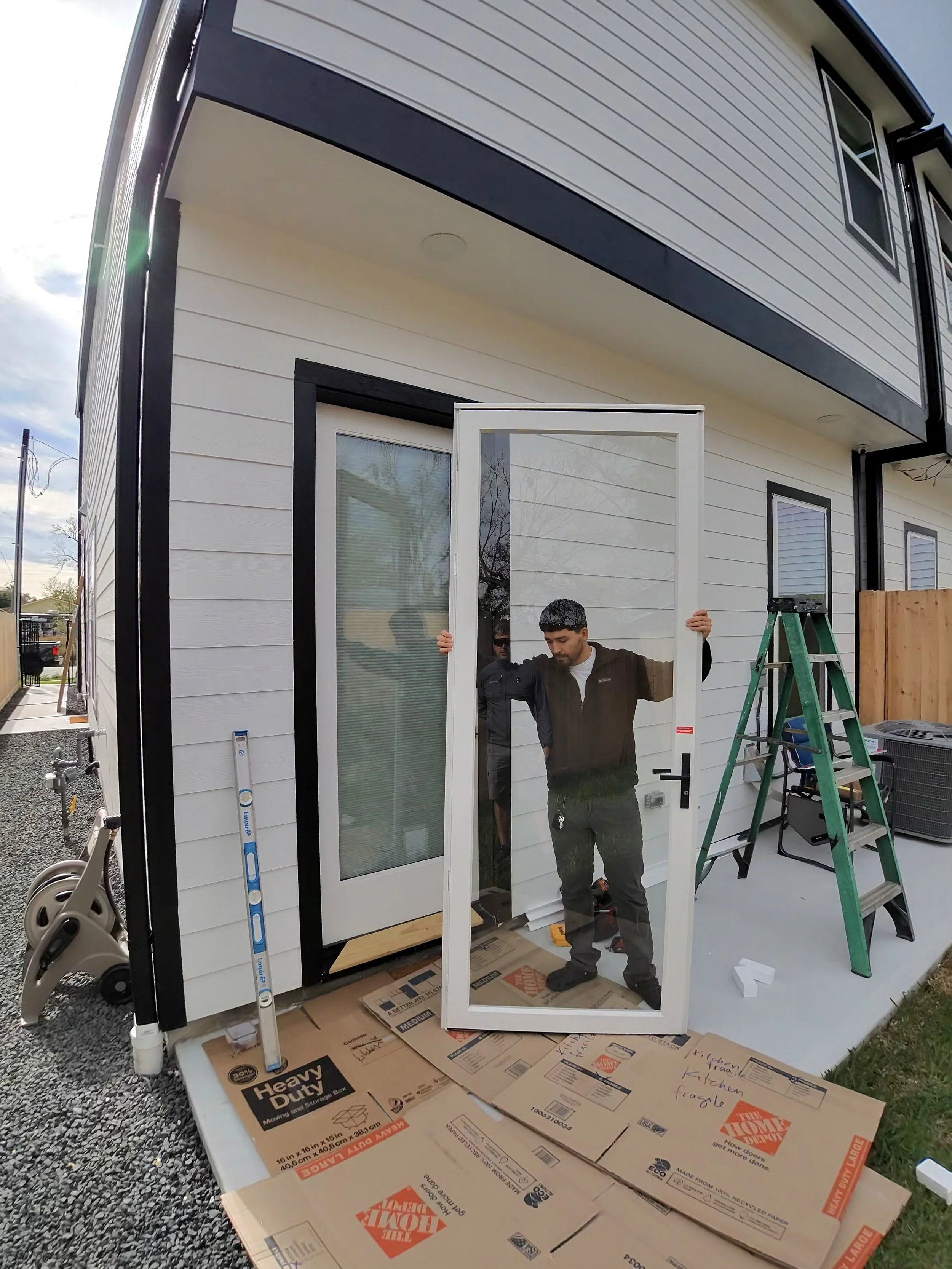 Man installing a glass storm door on a house exterior, surrounded by packing boxes and a ladder.