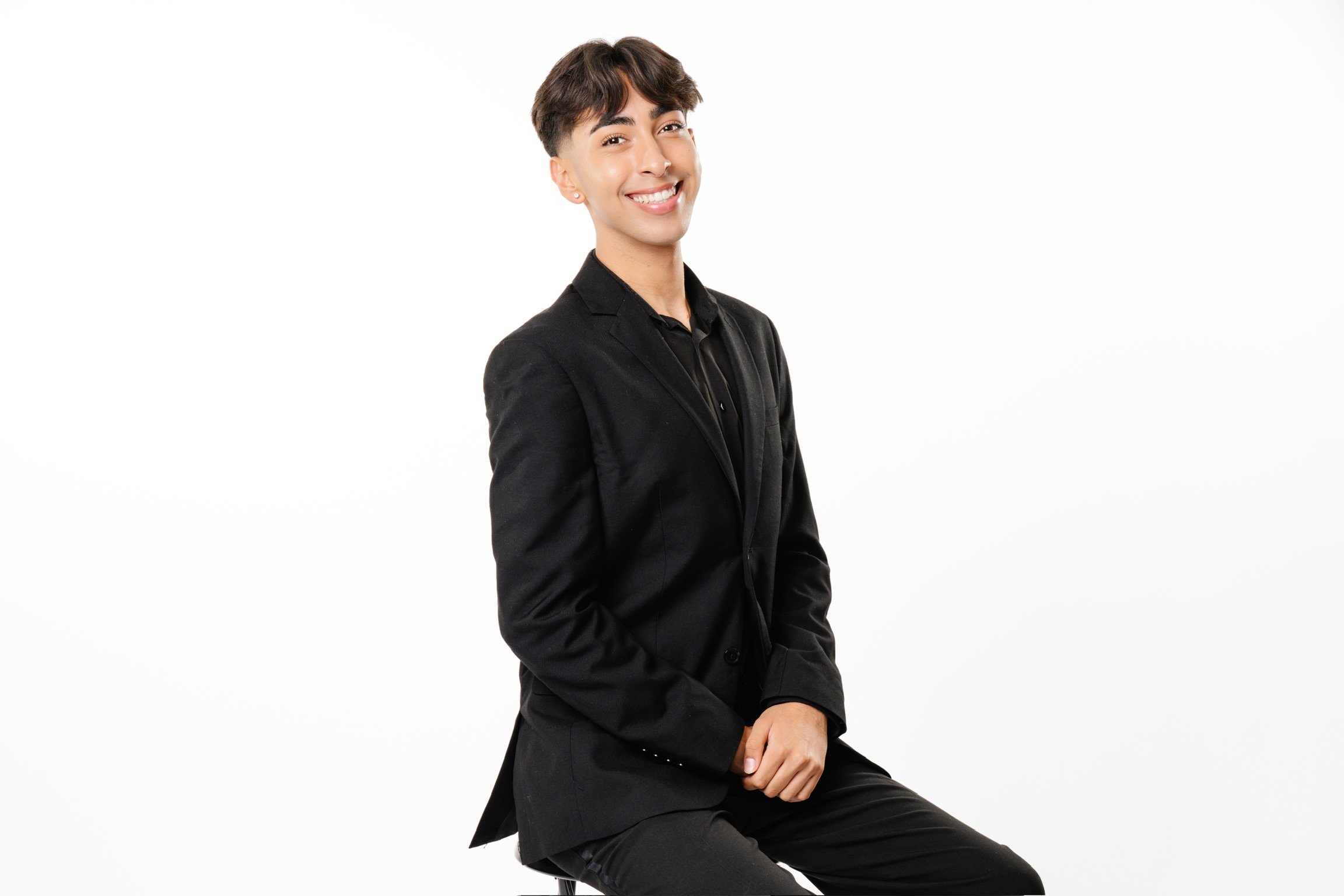 A young person with short brown hair wearing a black blazer and black shirt, smiling and sitting on a stool against a plain white background.