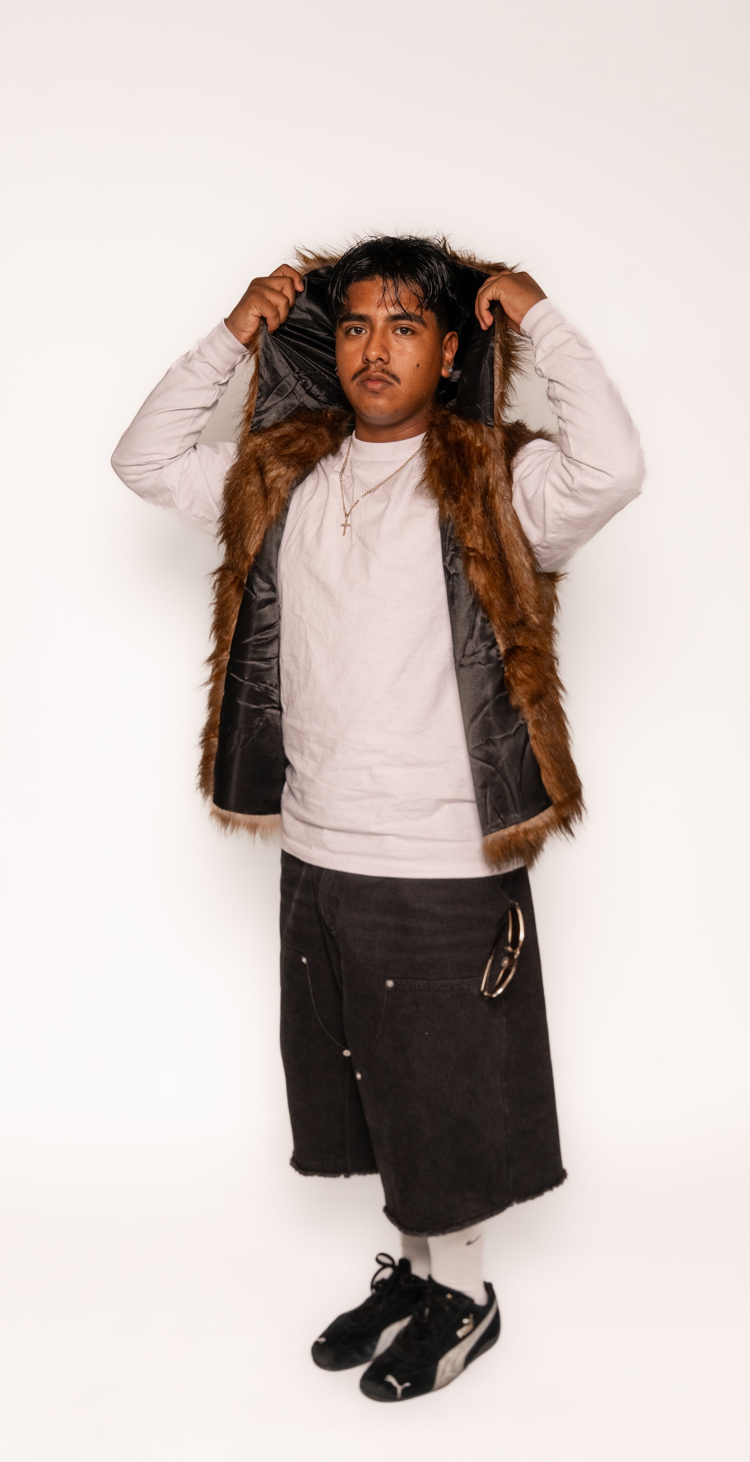 A person with dark hair, wearing a gold cross necklace, a gray shirt, a faux fur-lined vest, black shorts, and black sneakers, standing against a plain white background.
