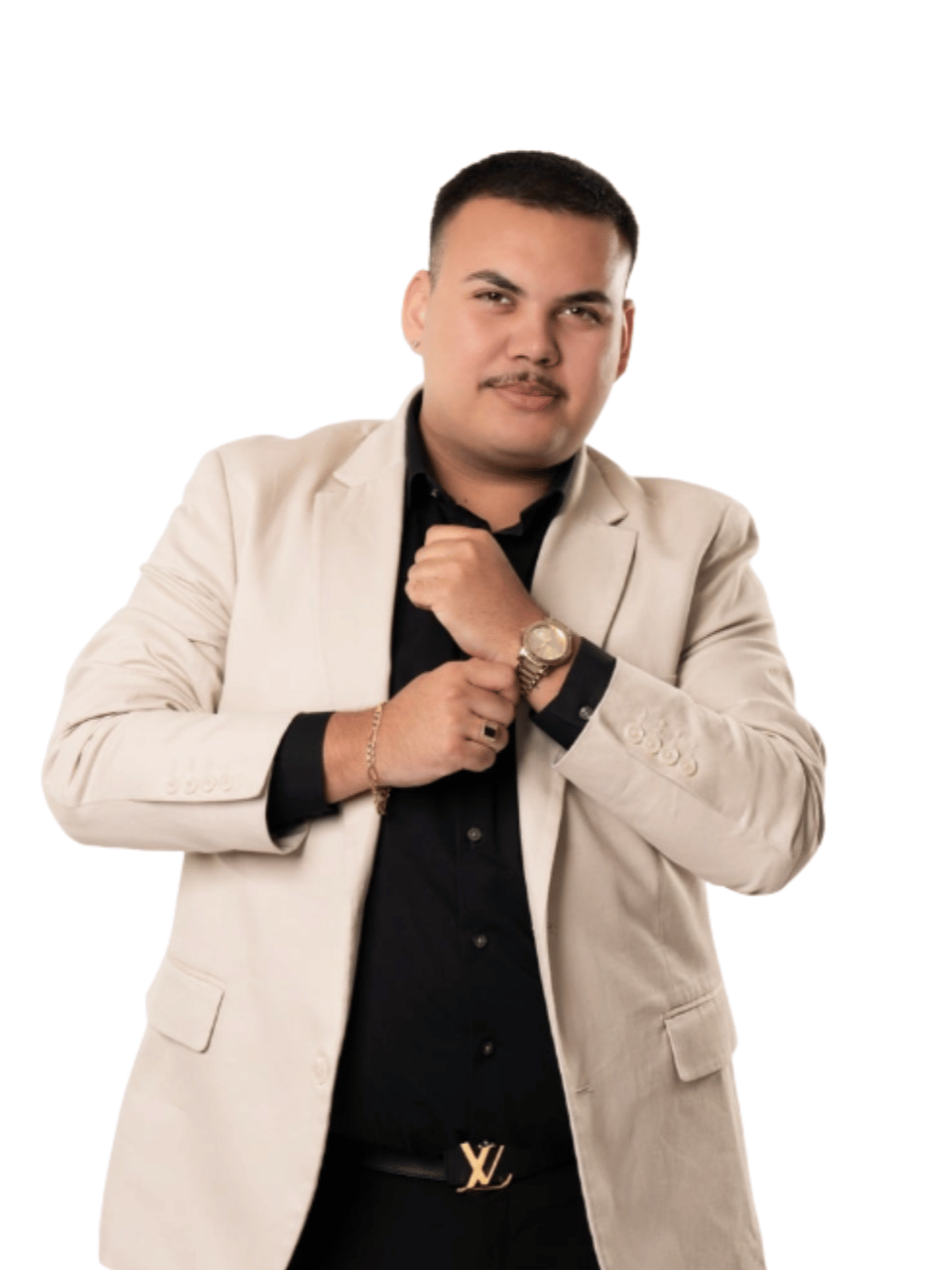 A young man with short dark hair, wearing a beige blazer over a black shirt, adjusting his blazer with one hand and looking confidently at the camera against a white background.