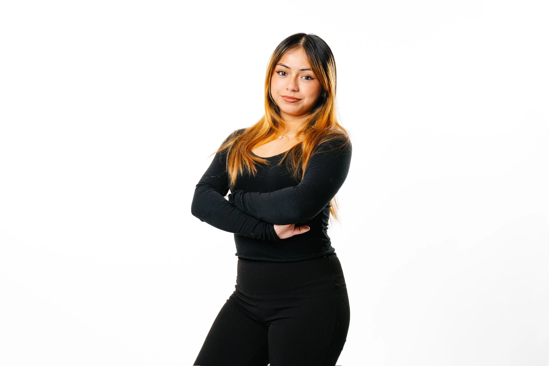 A young woman with long, wavy hair wearing a black long-sleeve top and black pants, standing with her arms crossed in front of a white background.