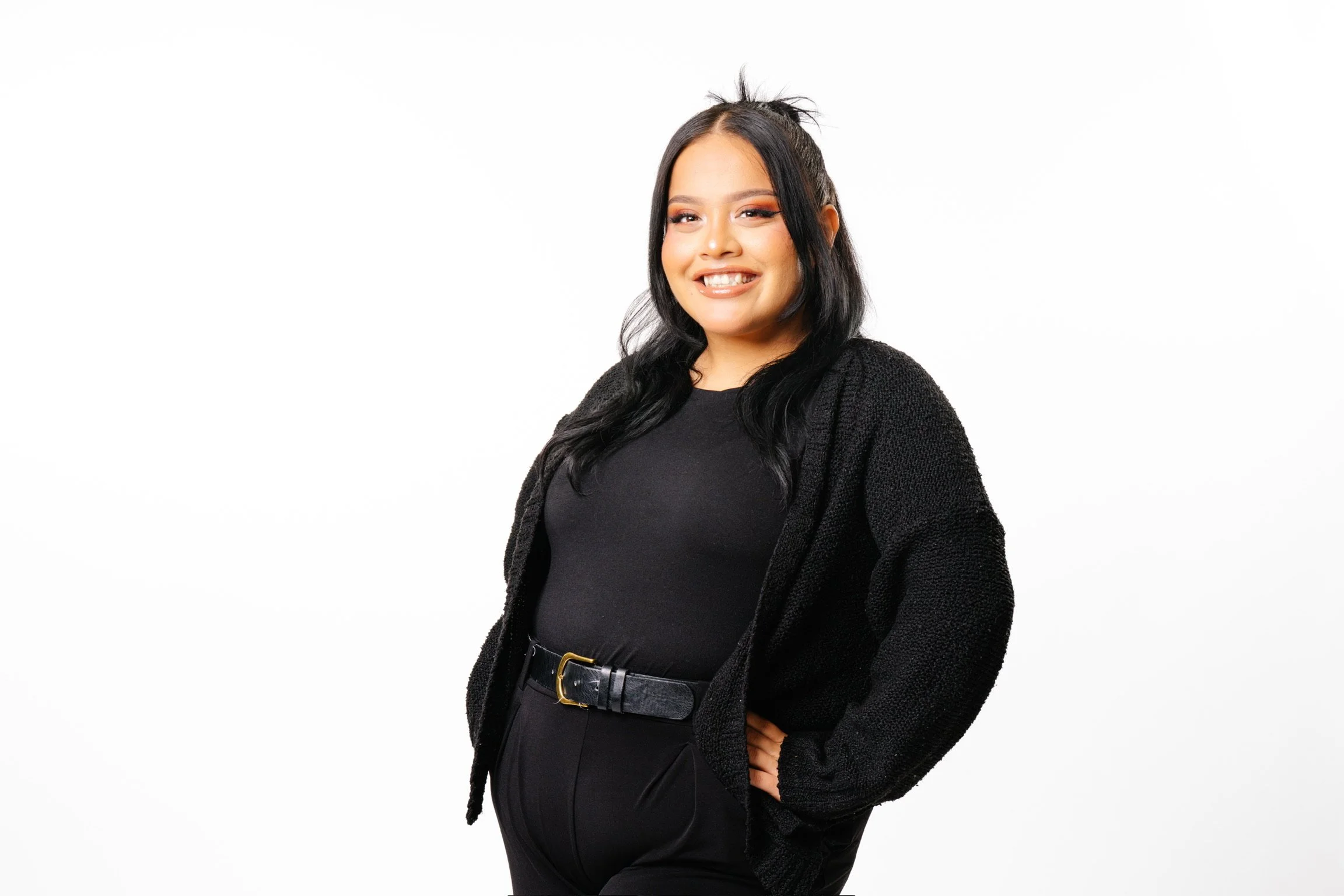 A young woman with long black hair styled with a half-up bun, wearing a black top, black cardigan, and high-waisted black pants with a belt, smiling with her hand on her hip against a white background.