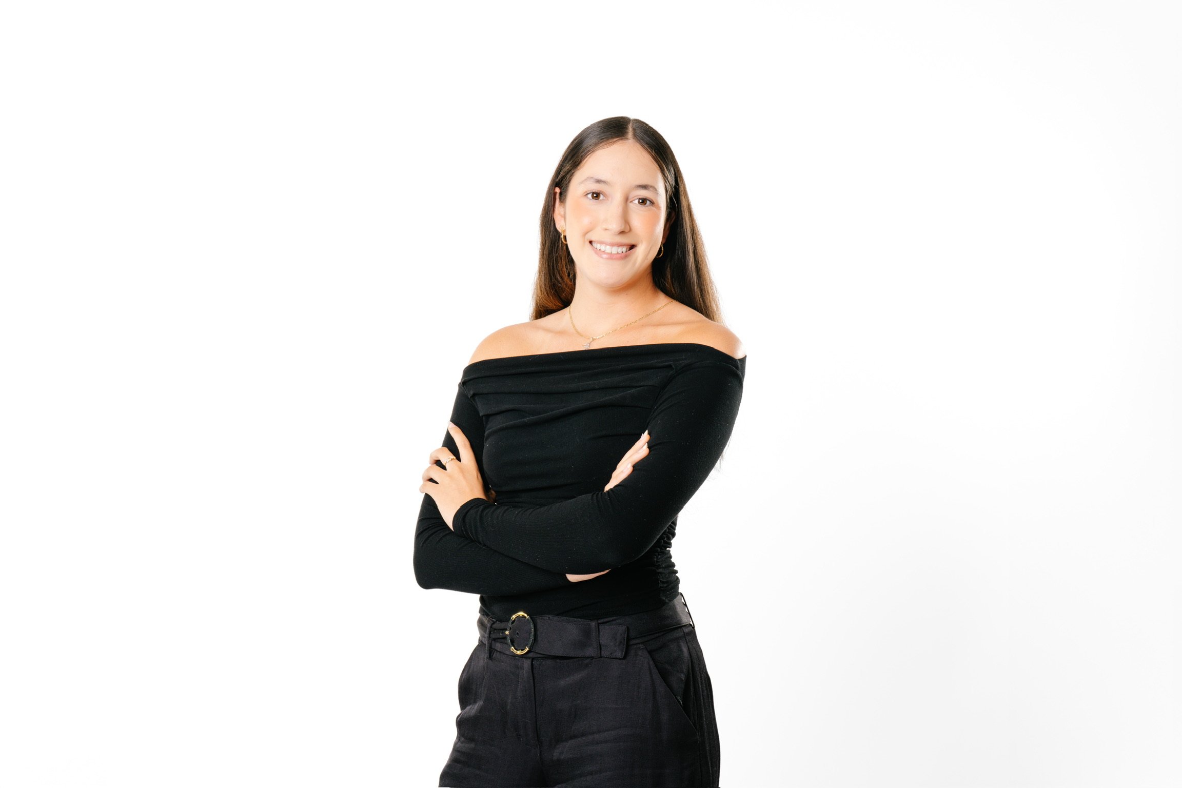 A young woman with long brown hair, smiling, wearing a black off-the-shoulder top with long sleeves, black pants with a belt, standing with arms crossed against a plain white background.