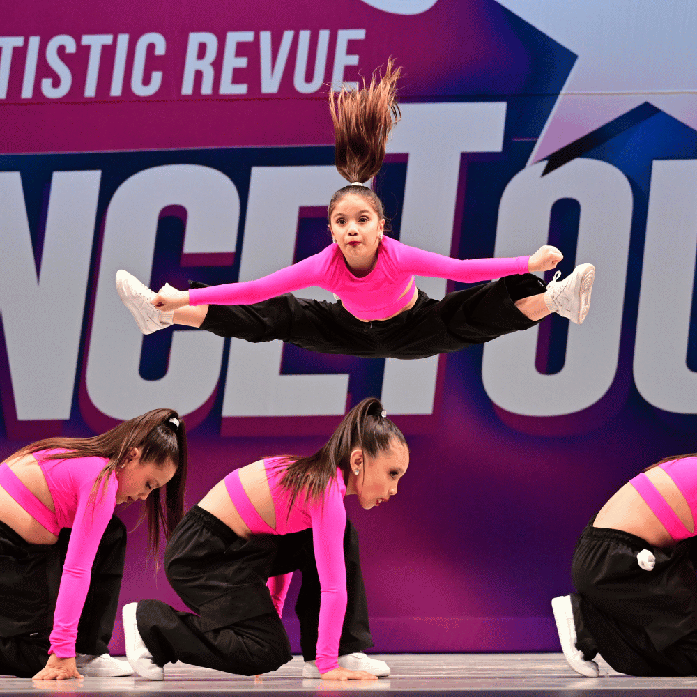 Four young female dancers in pink tops and black pants performing a dance routine on stage. One dancer is mid-air doing a leap, while the other three are kneeling or crouching on the floor.