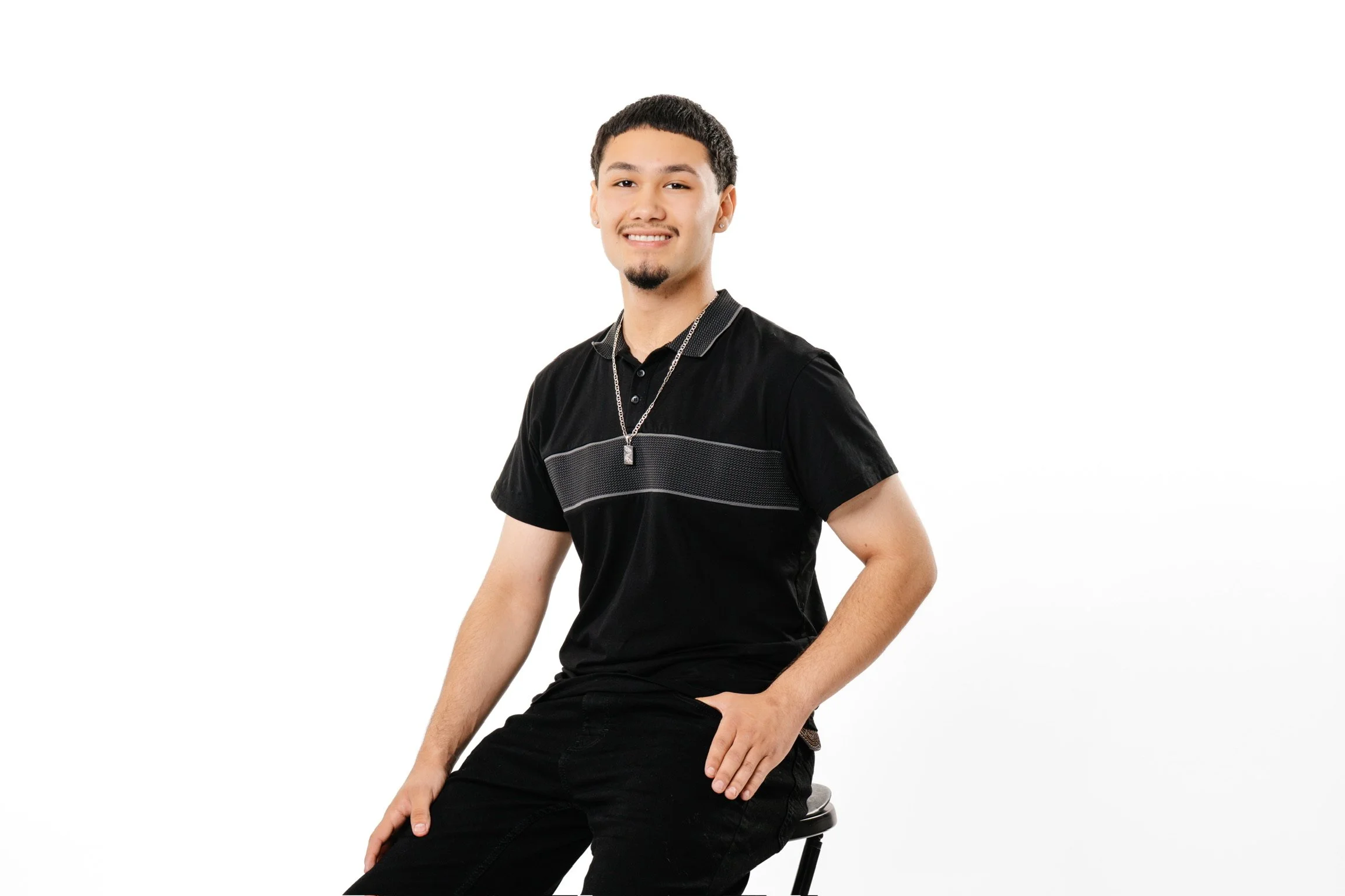 A young man with short black hair and a goatee, sitting on a black stool, smiling slightly and wearing a black polo shirt with a silver necklace, against a plain white background.