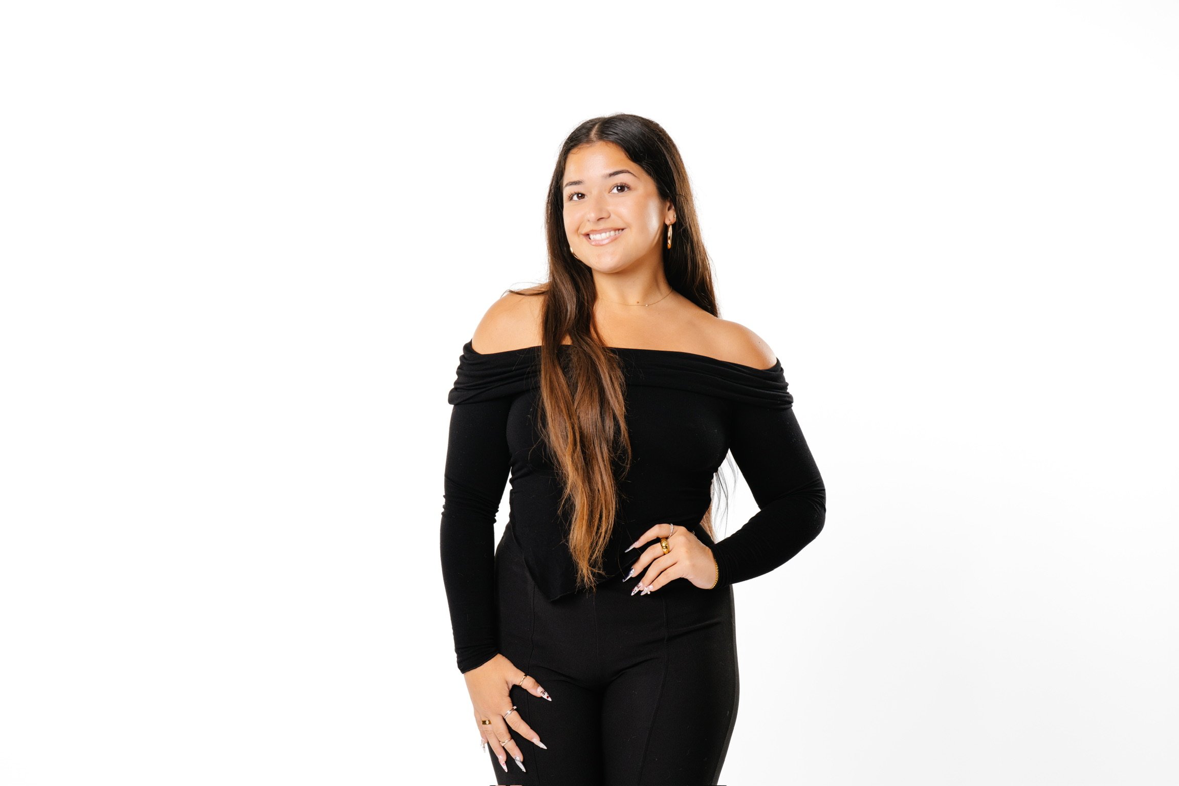 A young woman with long brunette hair in an off-the-shoulder black top and black pants, smiling and standing against a plain white background.