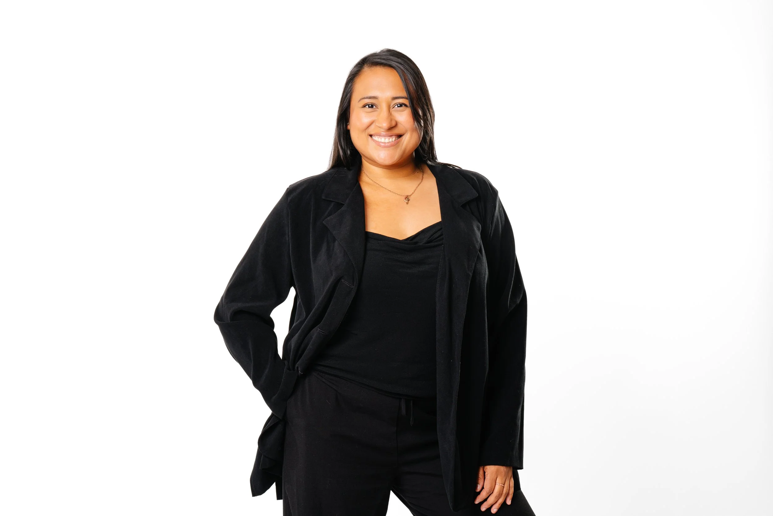 A woman with medium-length black hair smiling, wearing a black blazer and black top, standing against a white background.