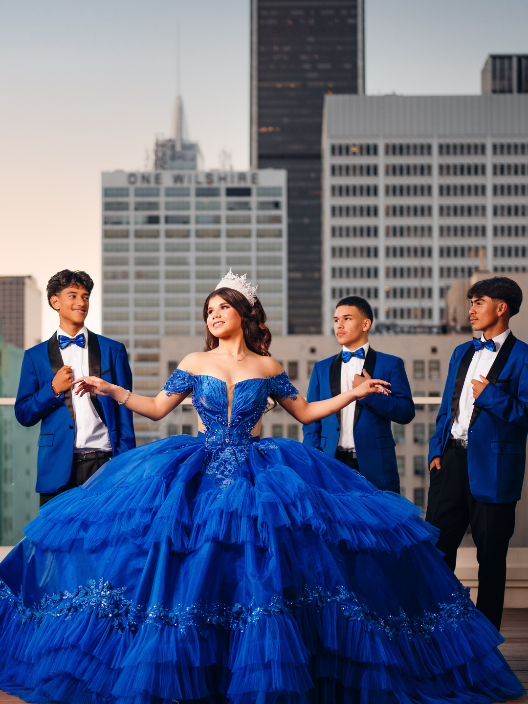 Young woman in an elaborate blue gown with a crown, surrounded by four young men in matching blue tuxedos, on a city rooftop with skyscrapers in the background.