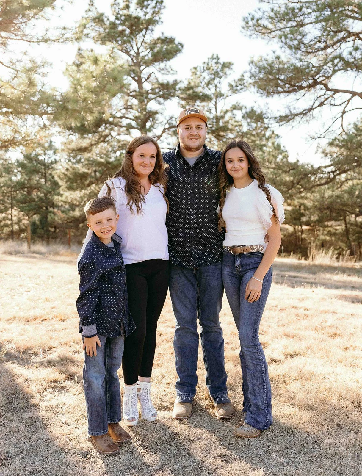 Family of five standing outdoors in a park or field with trees in the background, smiling at the camera.