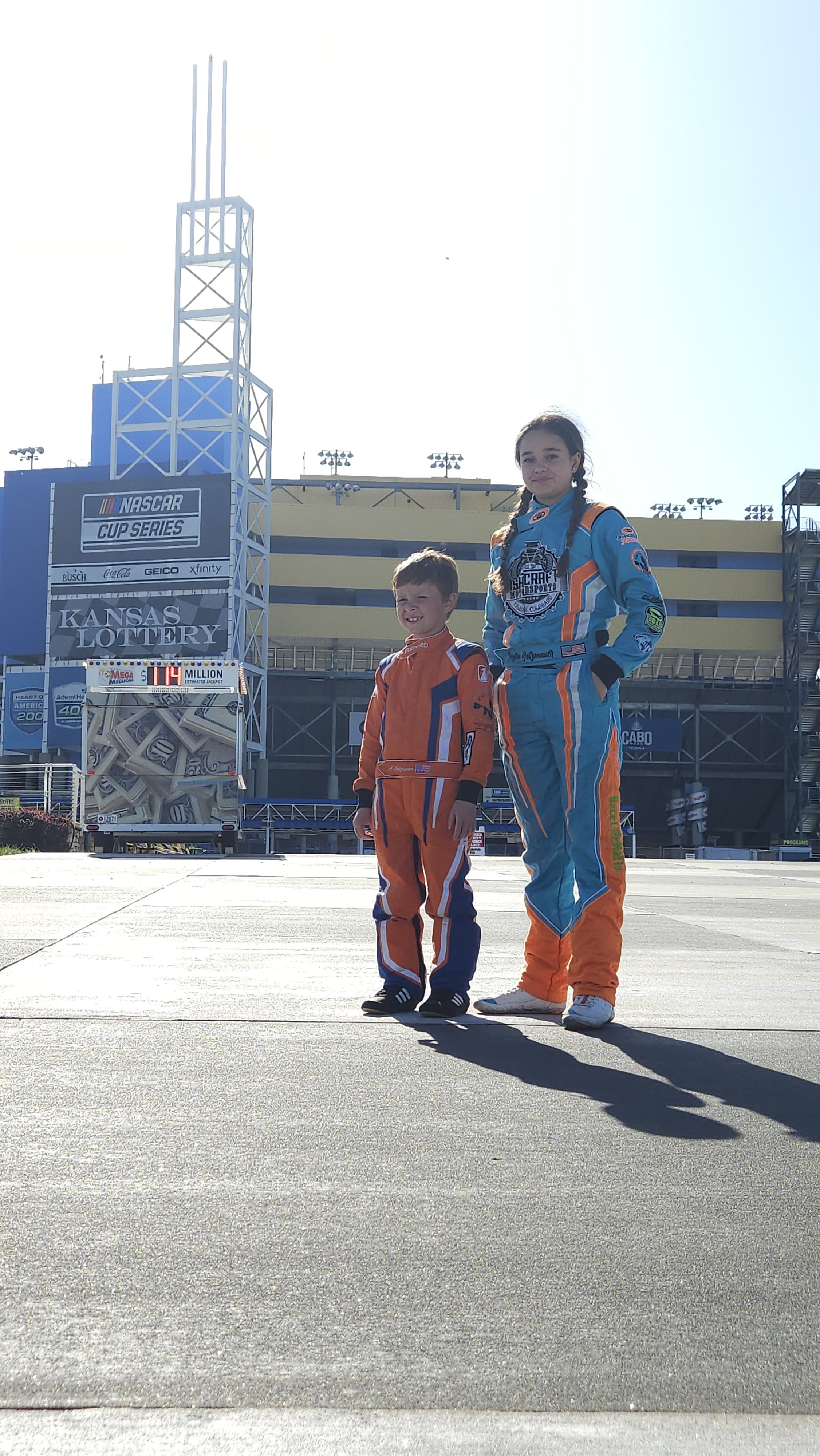 Two young children, a girl and a boy, in racing suits standing outside near a race track with a scoreboard and large building in the background.