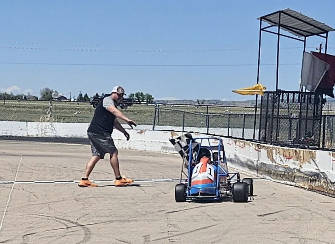 A person in casual clothing and a hat holding a flag stands next to a small race car on a dirt track, with a fence and open field in the background.