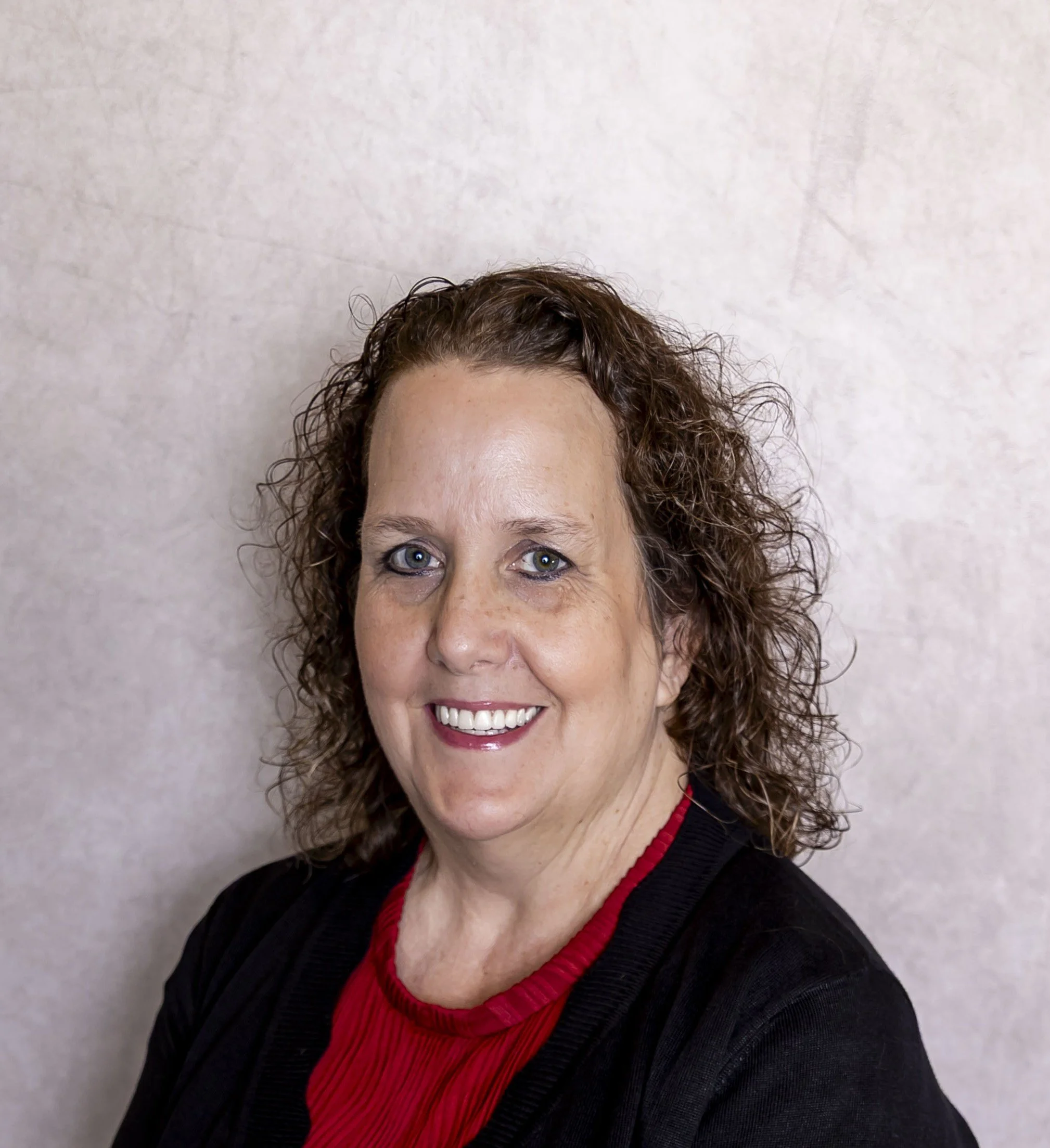 A woman with curly brown hair and blue eyes smiling, wearing a black and red jacket, in front of a light-colored textured wall.