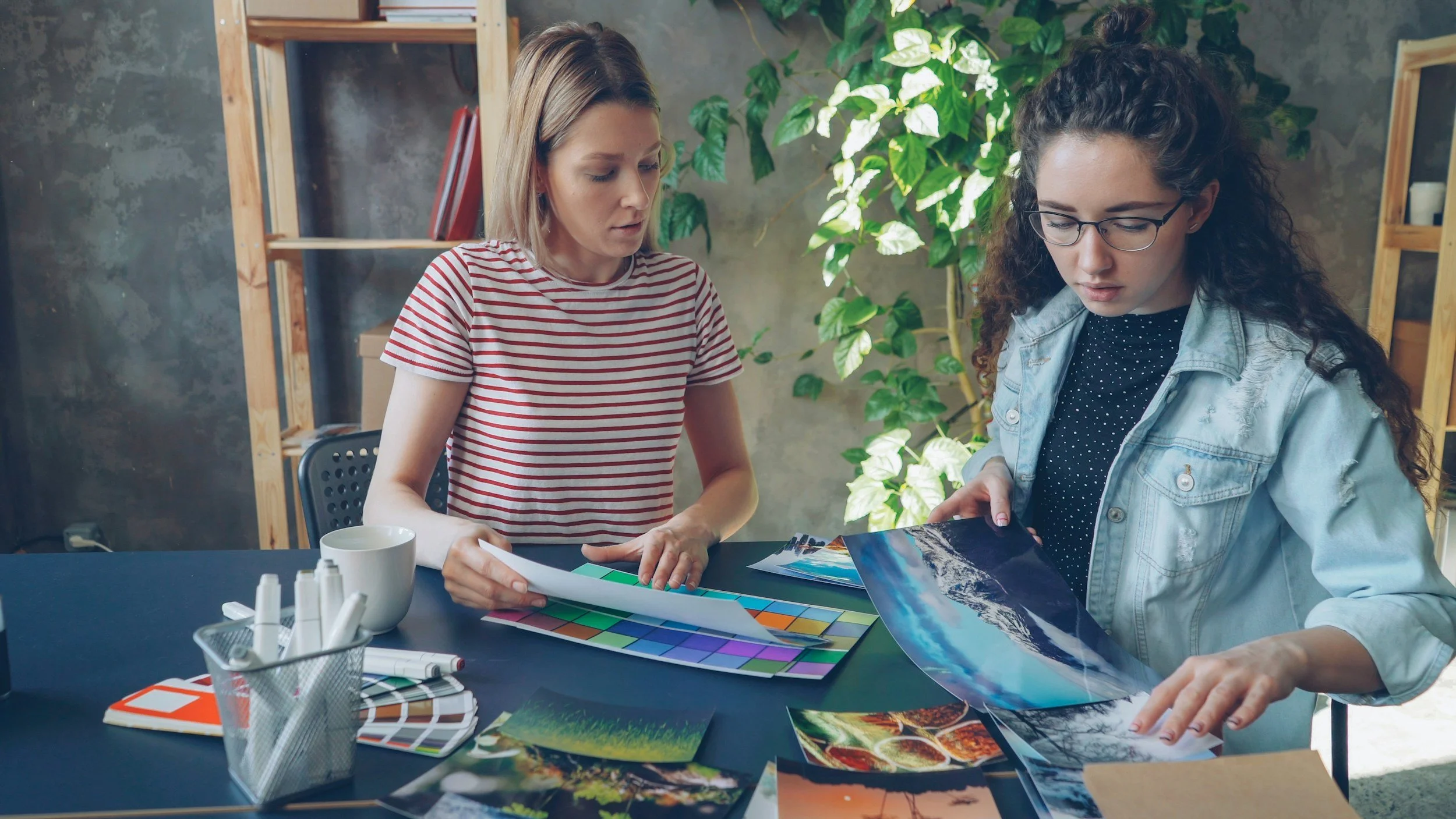 Two women sitting at a table reviewing photos and color swatches, surrounded by art supplies and photos in a creative workspace with large green plants and wooden shelves in the background.