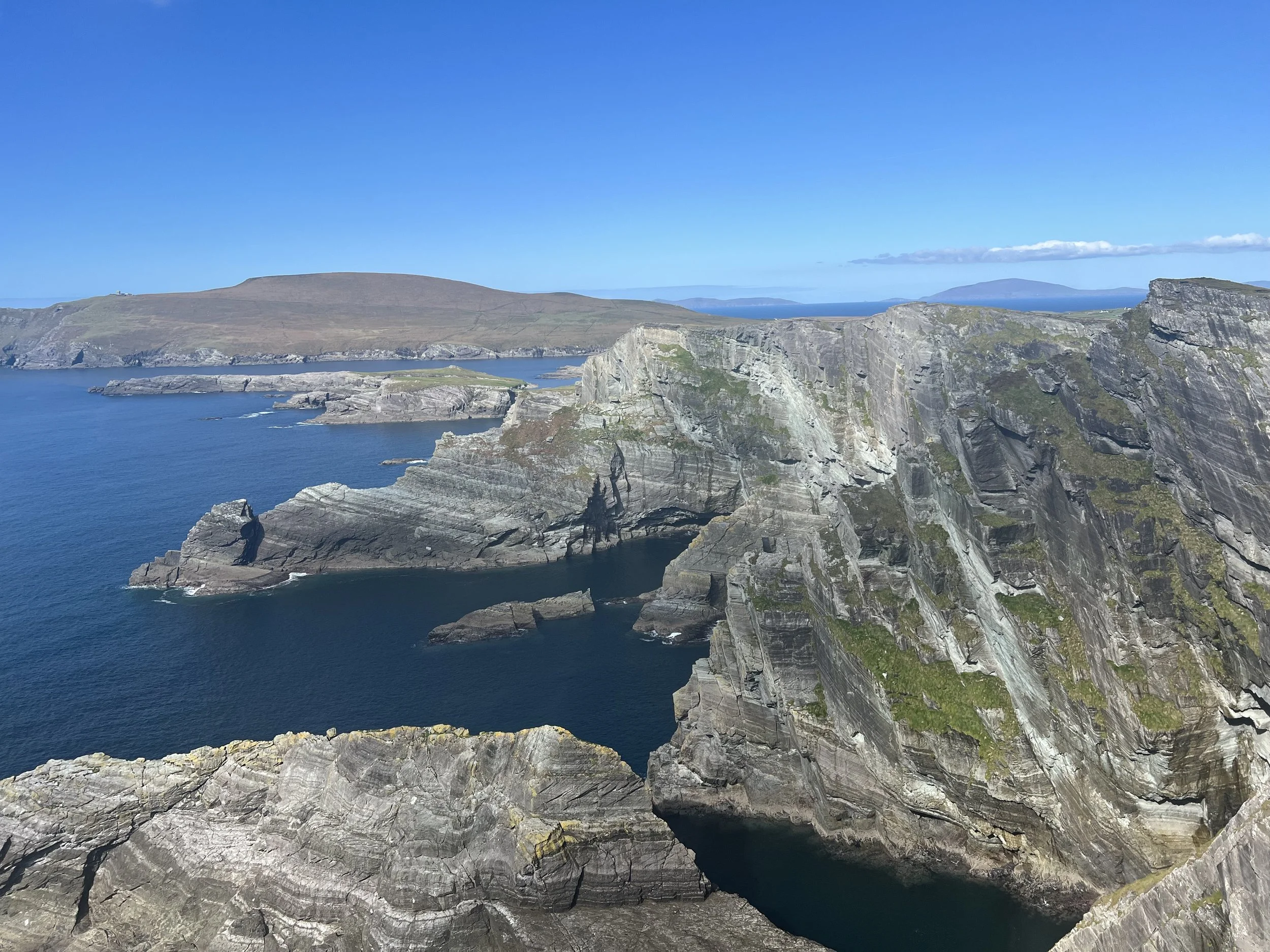 A scenic view of rocky cliffs overlooking the ocean with a hill in the background under a clear blue sky in Ireland