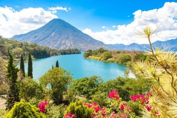 Scenic view of a lake with lush greenery, flowering plants, and a mountain in the background under a partly cloudy sky. Guatemala