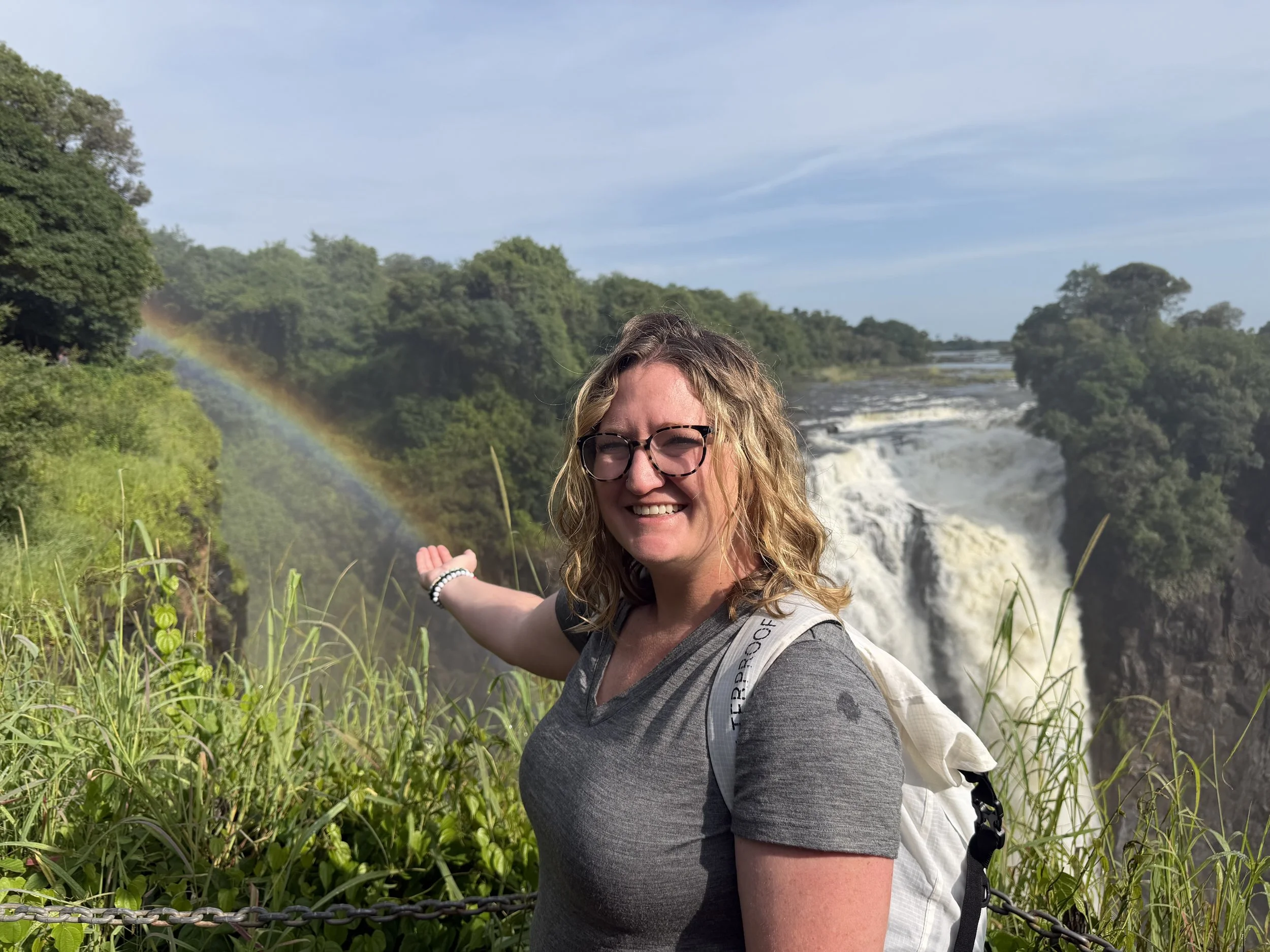 A woman with glasses and wavy blonde hair standing outdoors near greenery and a waterfall with a rainbow in the background. Victoria Falls