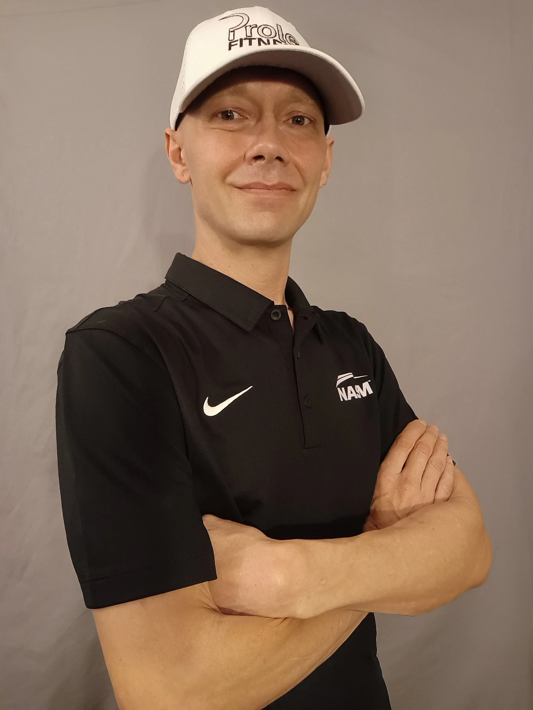 A man in a black Nike polo shirt and white baseball cap with logo that reads Prole Fitness, standing with arms crossed in front of a light gray background.