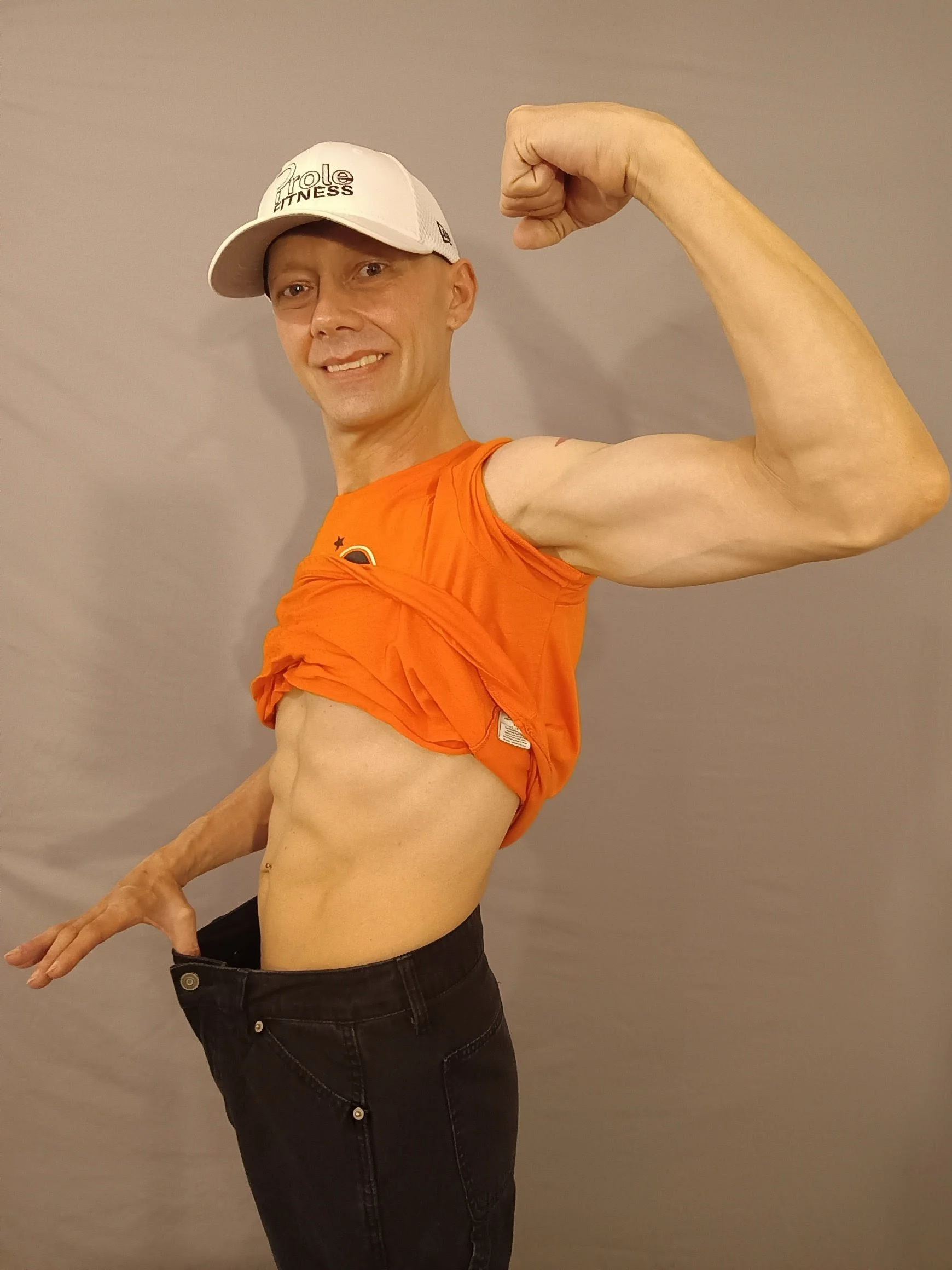 A man wearing a white cap and orange shirt flexes his bicep and points to his muscles with a big smile. He also wears black jeans and stands against a plain gray background.