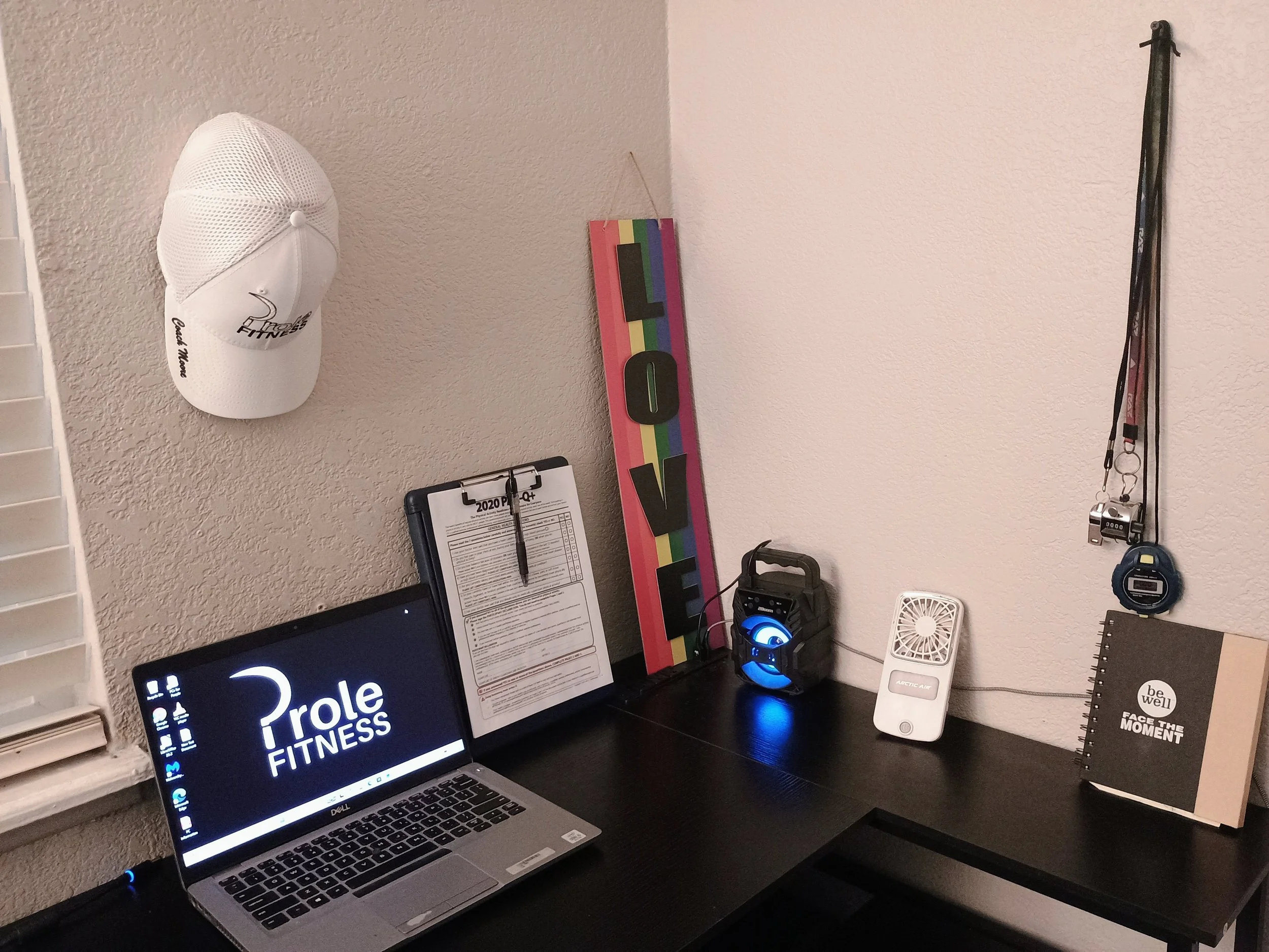 Home office desk with a laptop showing the Prole Fitness logo on the screen, a clipboard with papers, a rainbow-colored sign that says "LOVE," a white air purifier, a smartphone, a small fan, a water bottle, and workout accessories hanging on the wall.