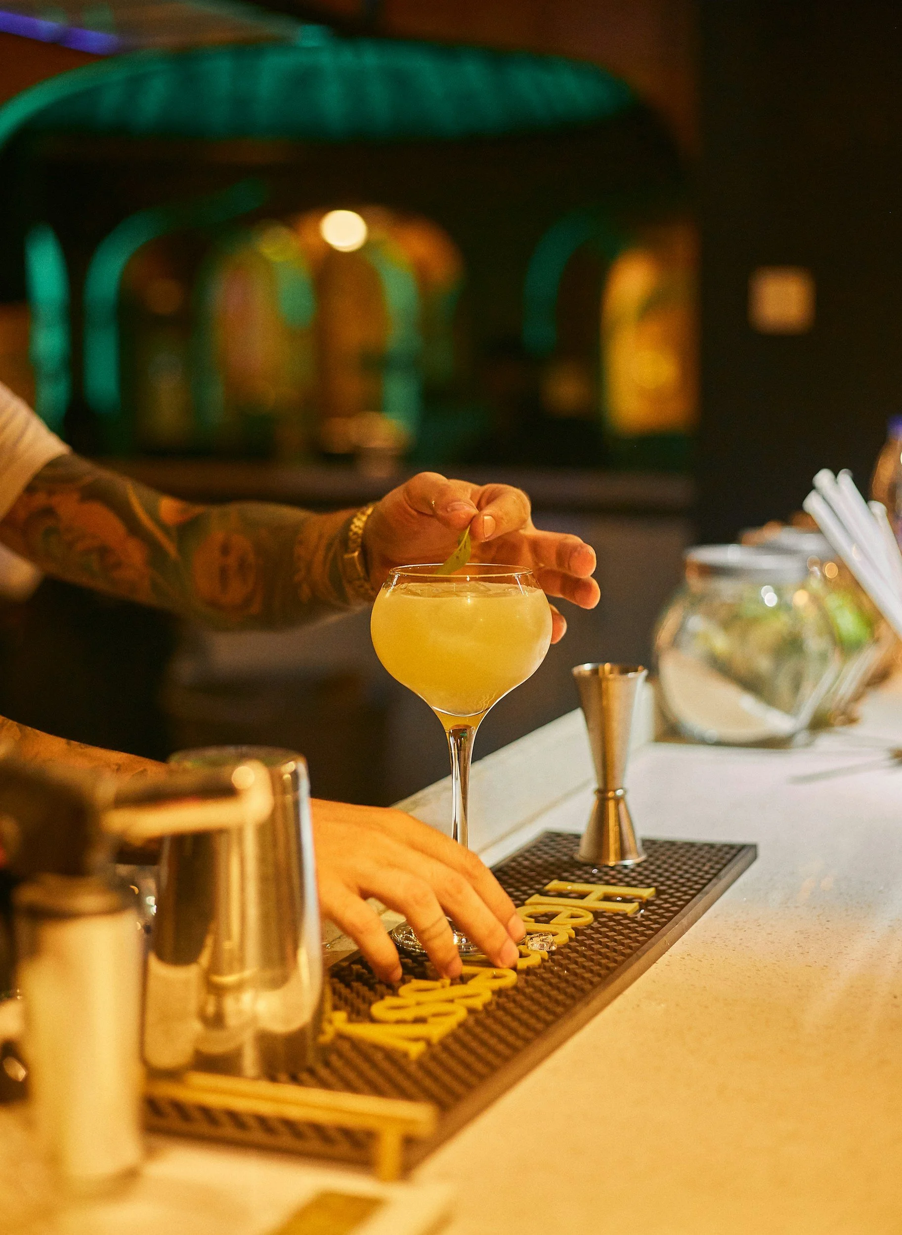 Person with tattoos stirring a cocktail at a bar counter with bar tools and garnishes, illuminated by dim lighting.