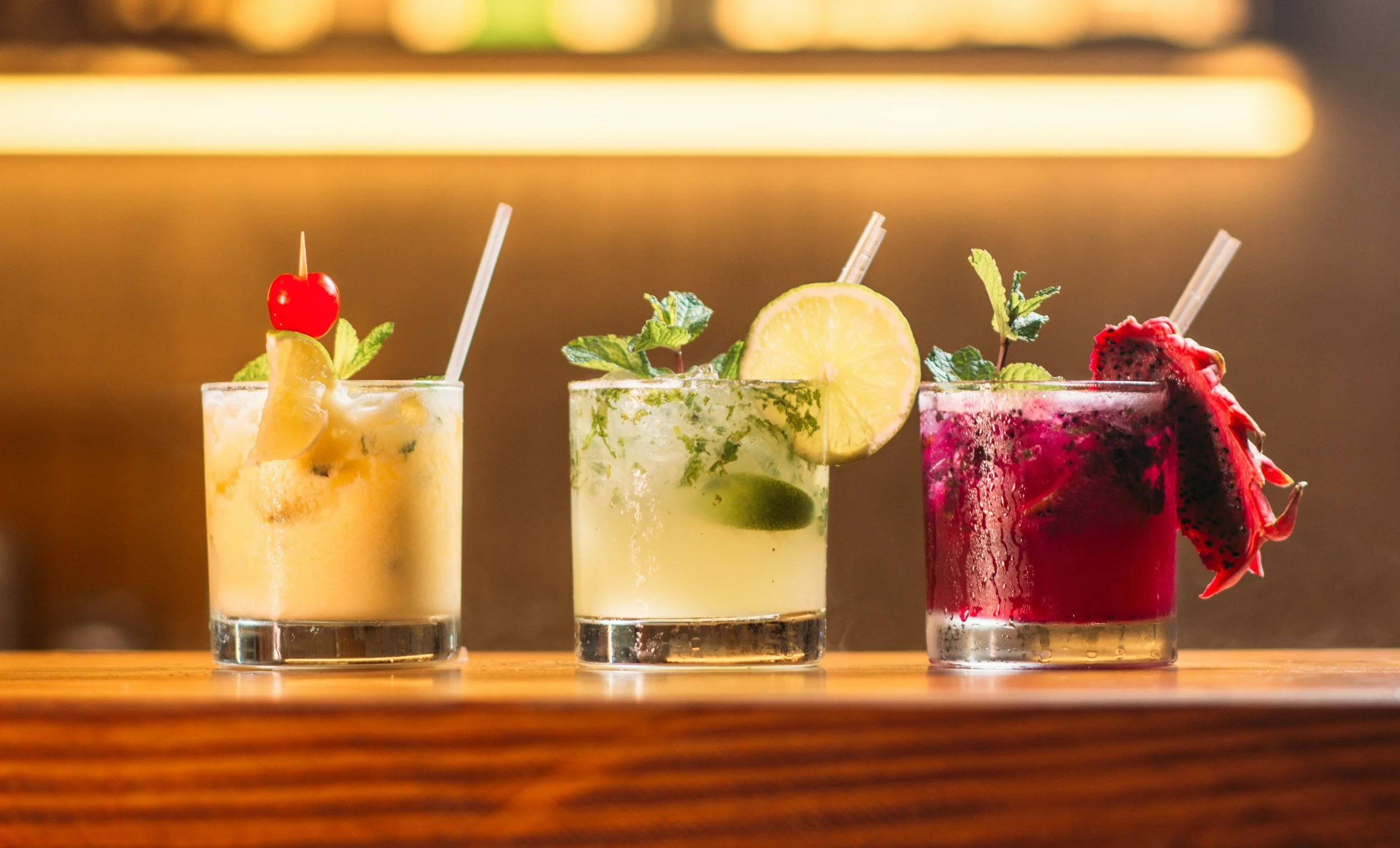 Three colorful cocktails garnished with fruit and herbs on a wooden surface.