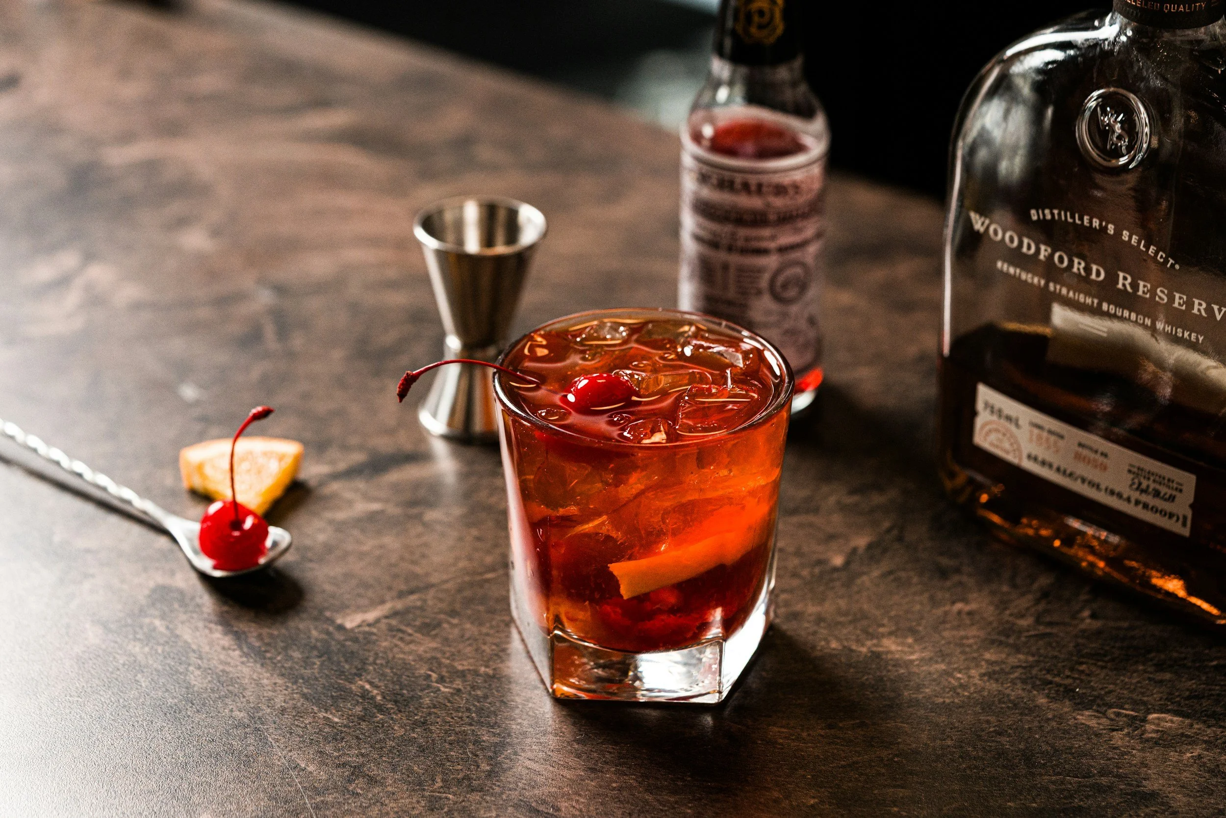 A glass of cocktail with ice and cherries on top, placed on a dark wooden surface. In the background, there is a bottle of Woodford Reserve bourbon whiskey, a small jar of bitters, and a metal jigger.
