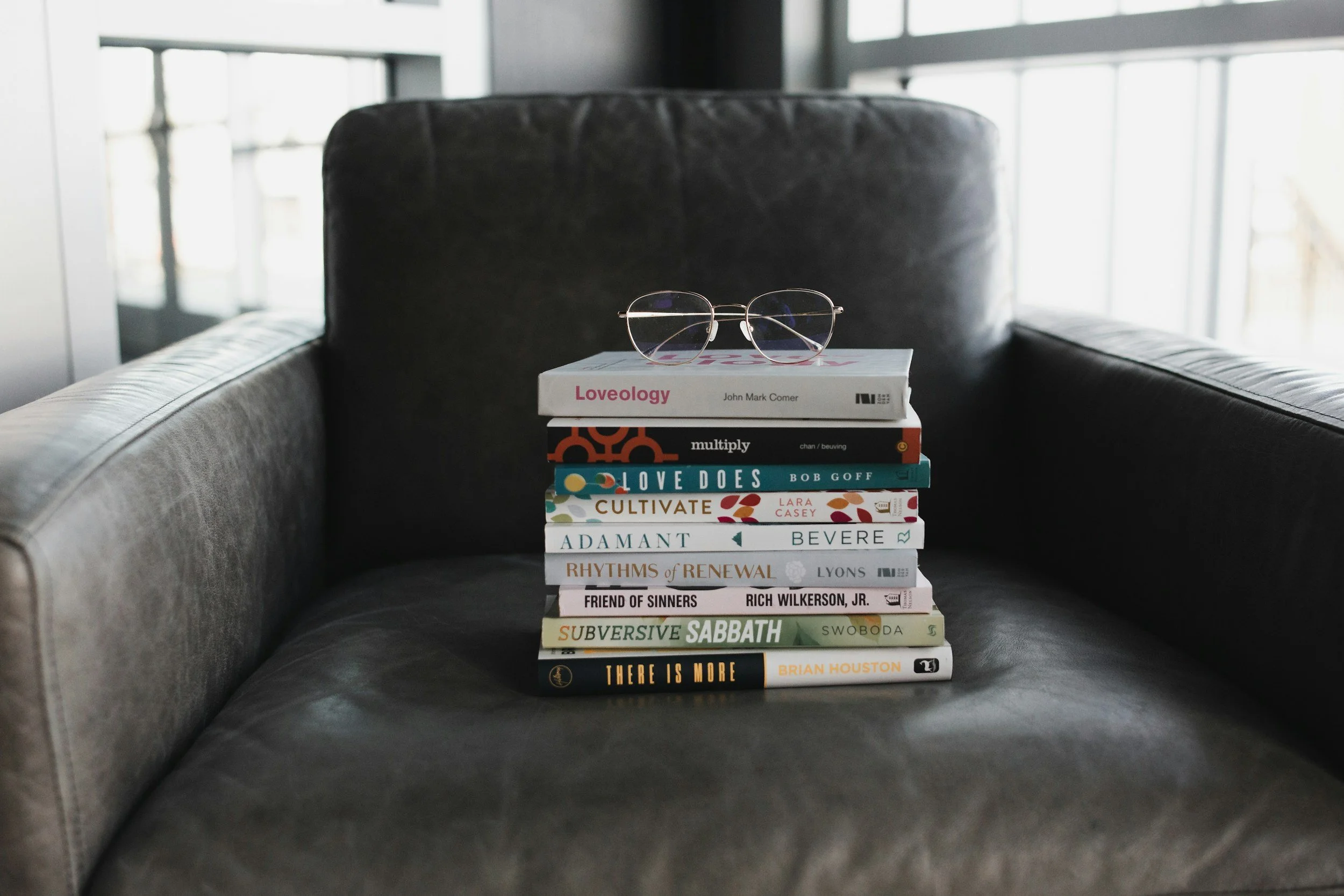 A stack of books on a dark gray couch with glasses on top, in front of large windows in a modern interior.