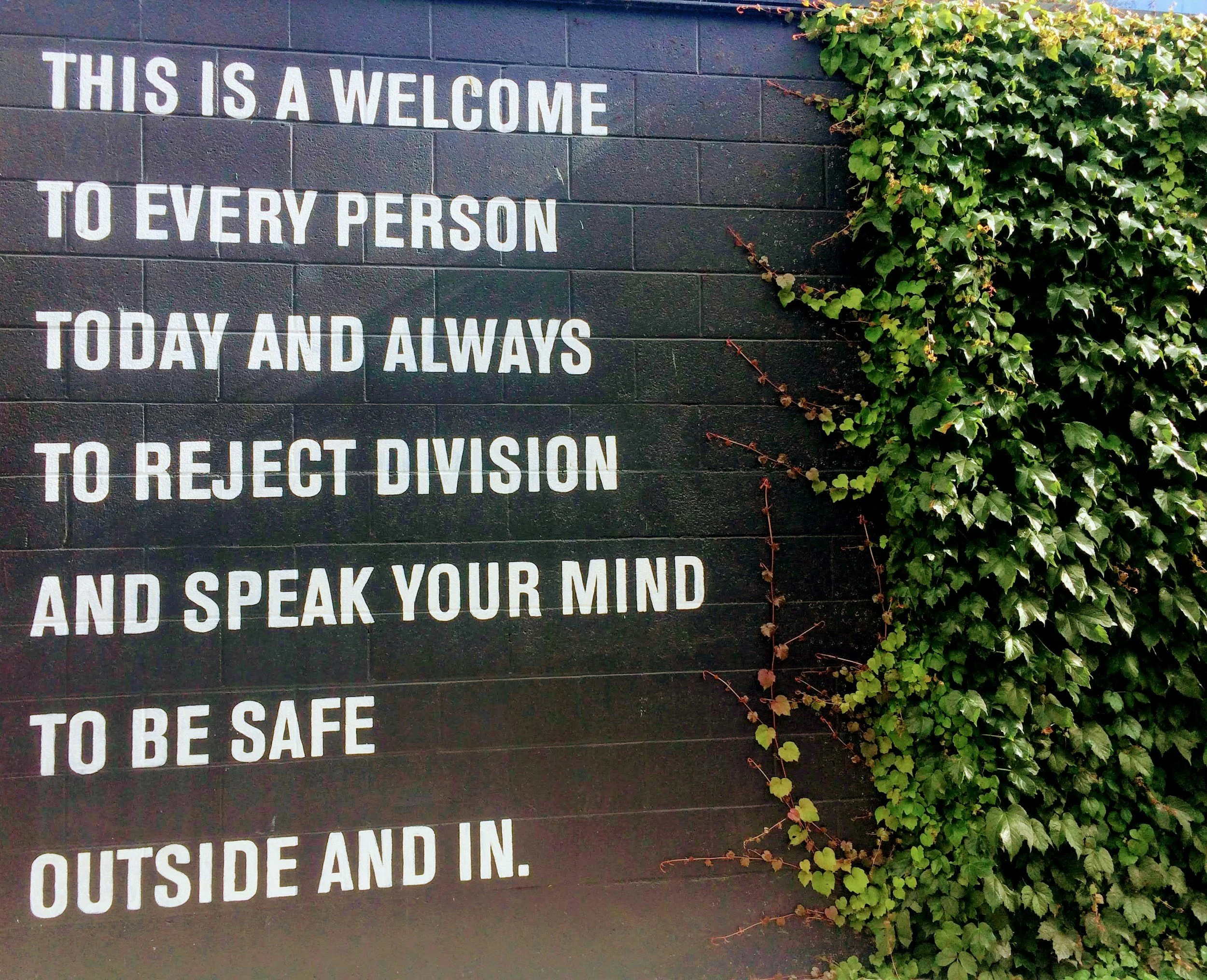 A black brick wall with white painted text that reads "This is a welcome to every person today and always to be rejected division outside and inside." The right side of the wall is covered with green ivy.